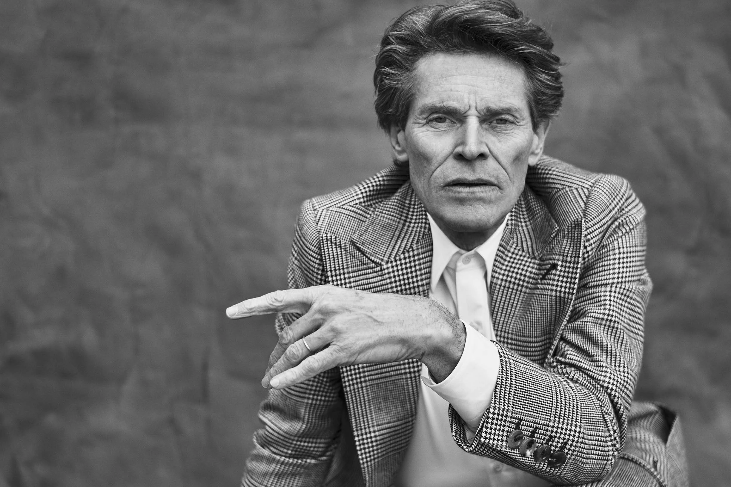 A black and white portrait of a man with wavy hair wearing a checkered blazer and a white shirt. He is looking directly at the camera with an intense expression and is pointing with his right hand.