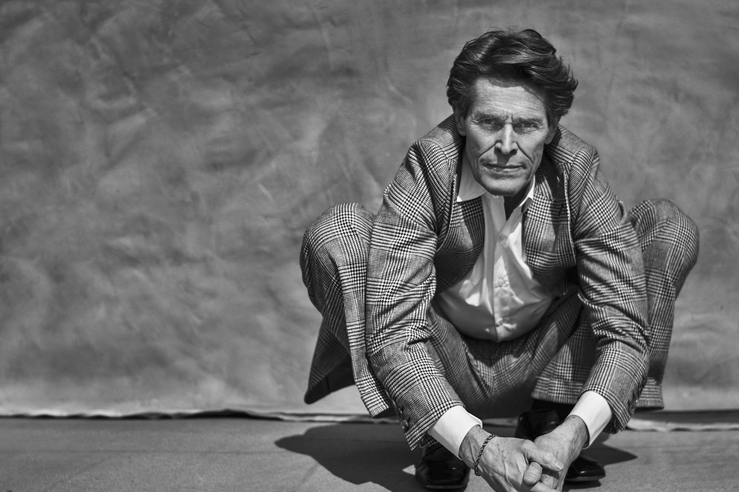 A man in a checkered suit squatting on the ground, looking directly at the camera, in black and white.