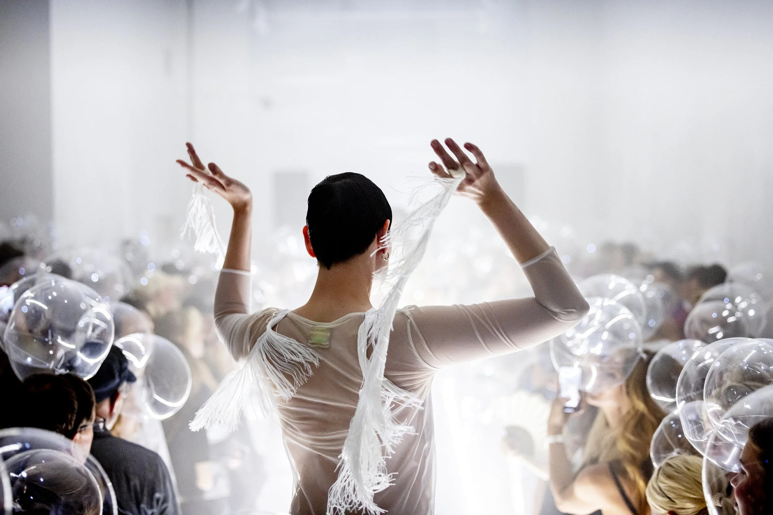 Person with short dark hair standing with back to the camera, arms raised, at a crowded event with balloons or inflated transparent spheres surrounding the crowd.