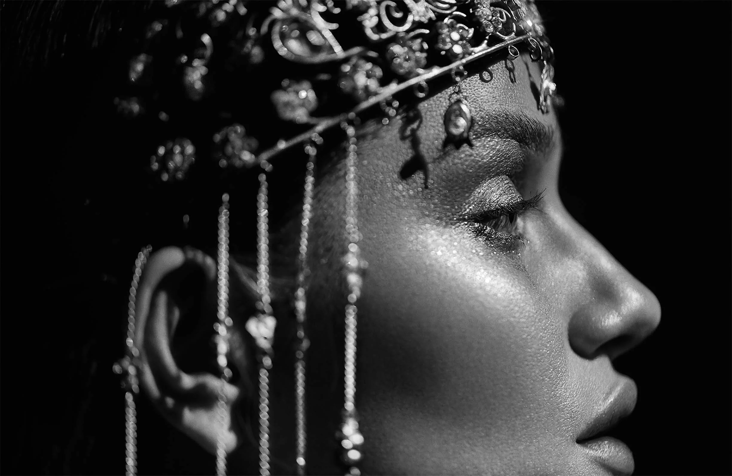 Close-up profile of a woman with textured skin, wearing an ornate headpiece with dangling chains and embellishments, in black and white.