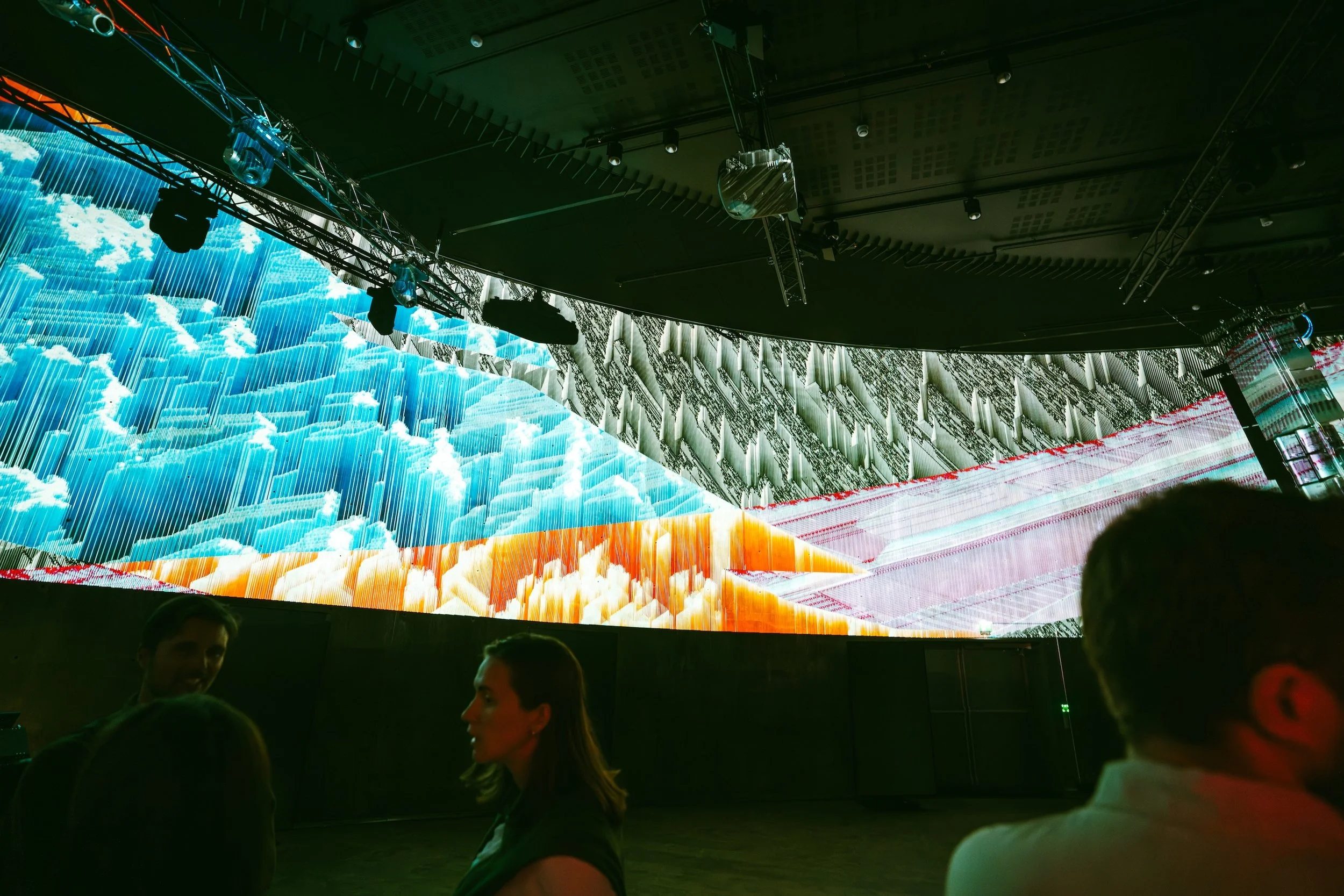 Large indoor screen displaying a digital image of clouds and rays of sunlight, with people observing in front of it.