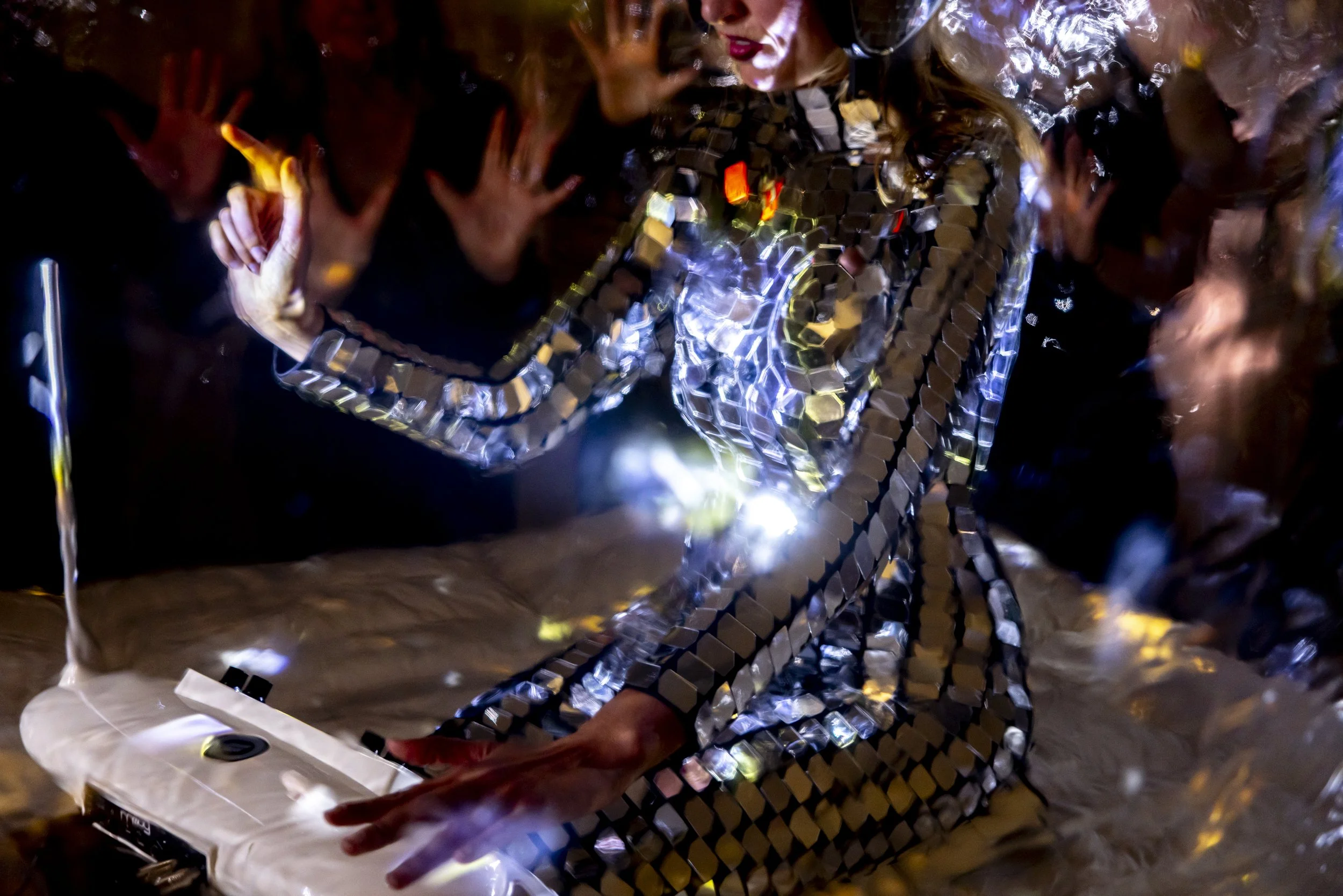 A woman in a reflective, mirrored outfit using a DJ mixer surrounded by a group of people, with some raising their hands and engaging in a dance or performance at a party or club.