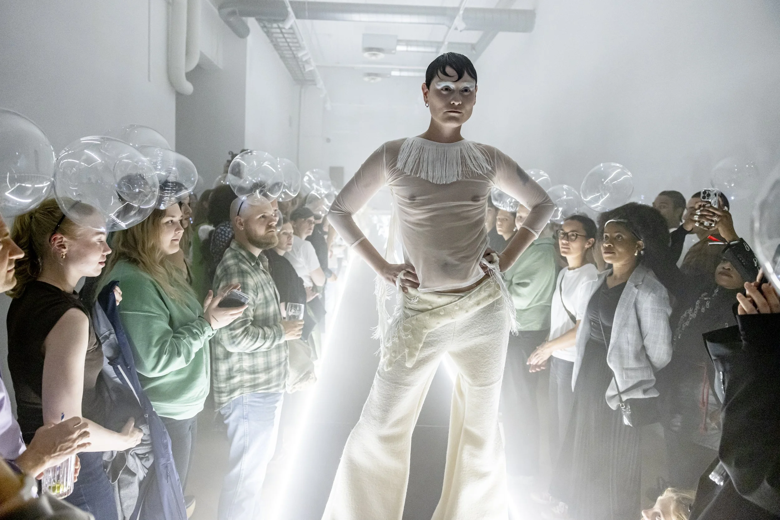 A tall person with white makeup and a unique hairstyle poses on a runway in front of an audience. The audience members are holding balloons or taking pictures, and the setting has a modern, industrial look with white walls and lighting.