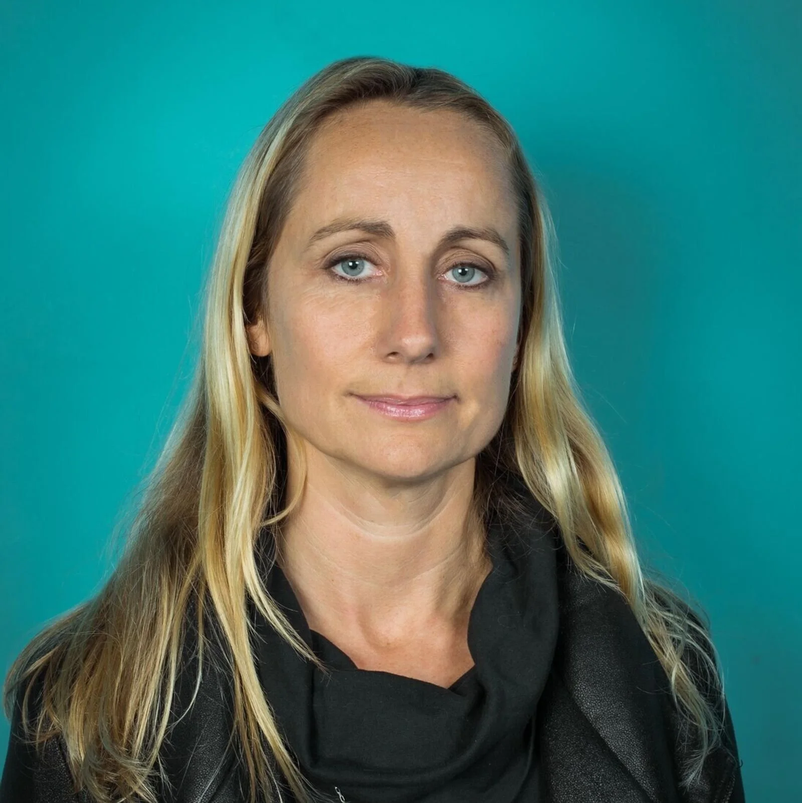 A woman with blonde hair and blue eyes wearing a black top and standing against a teal background.