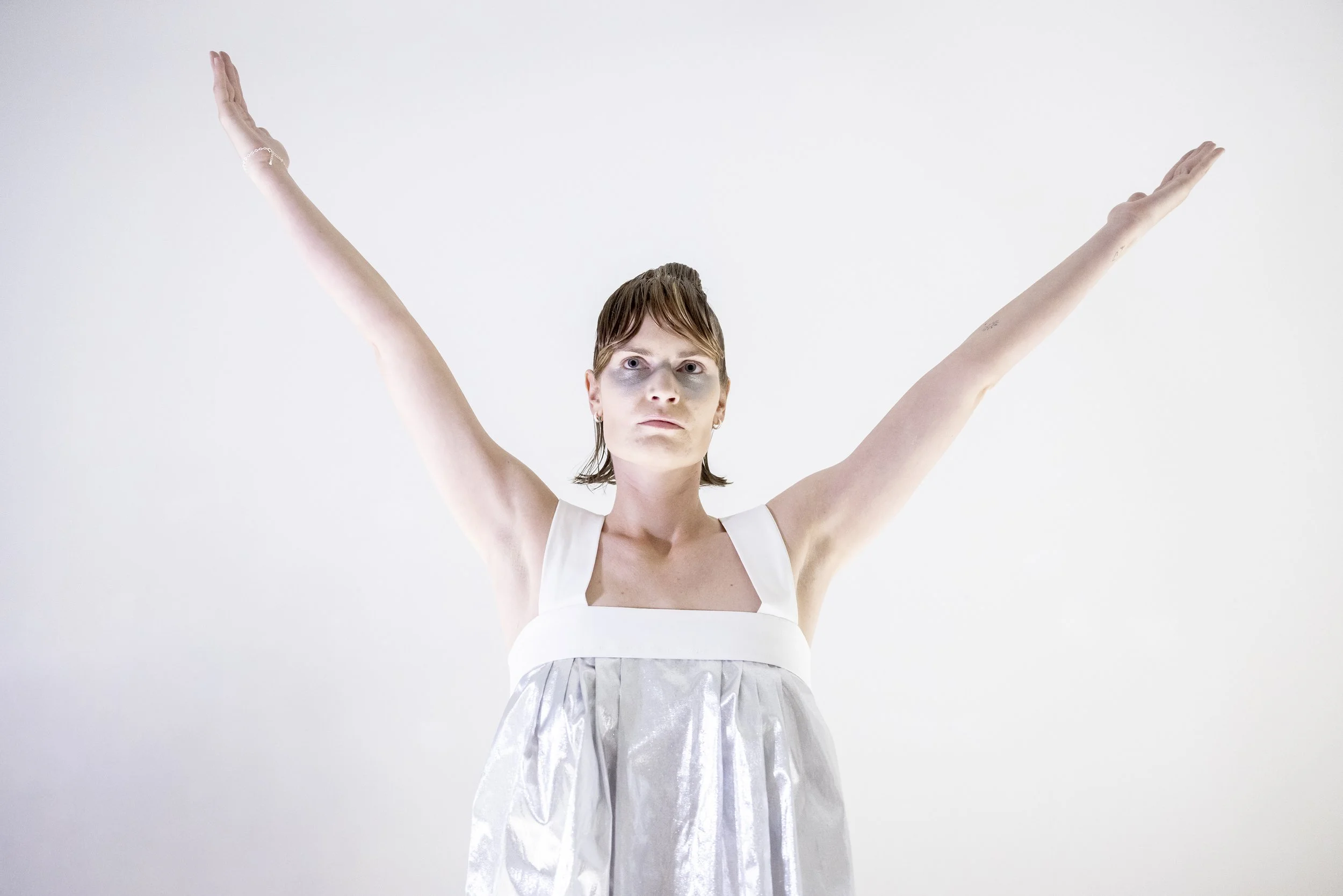 A woman with short brown hair and makeup standing against a plain white background with her arms raised and outstretched.