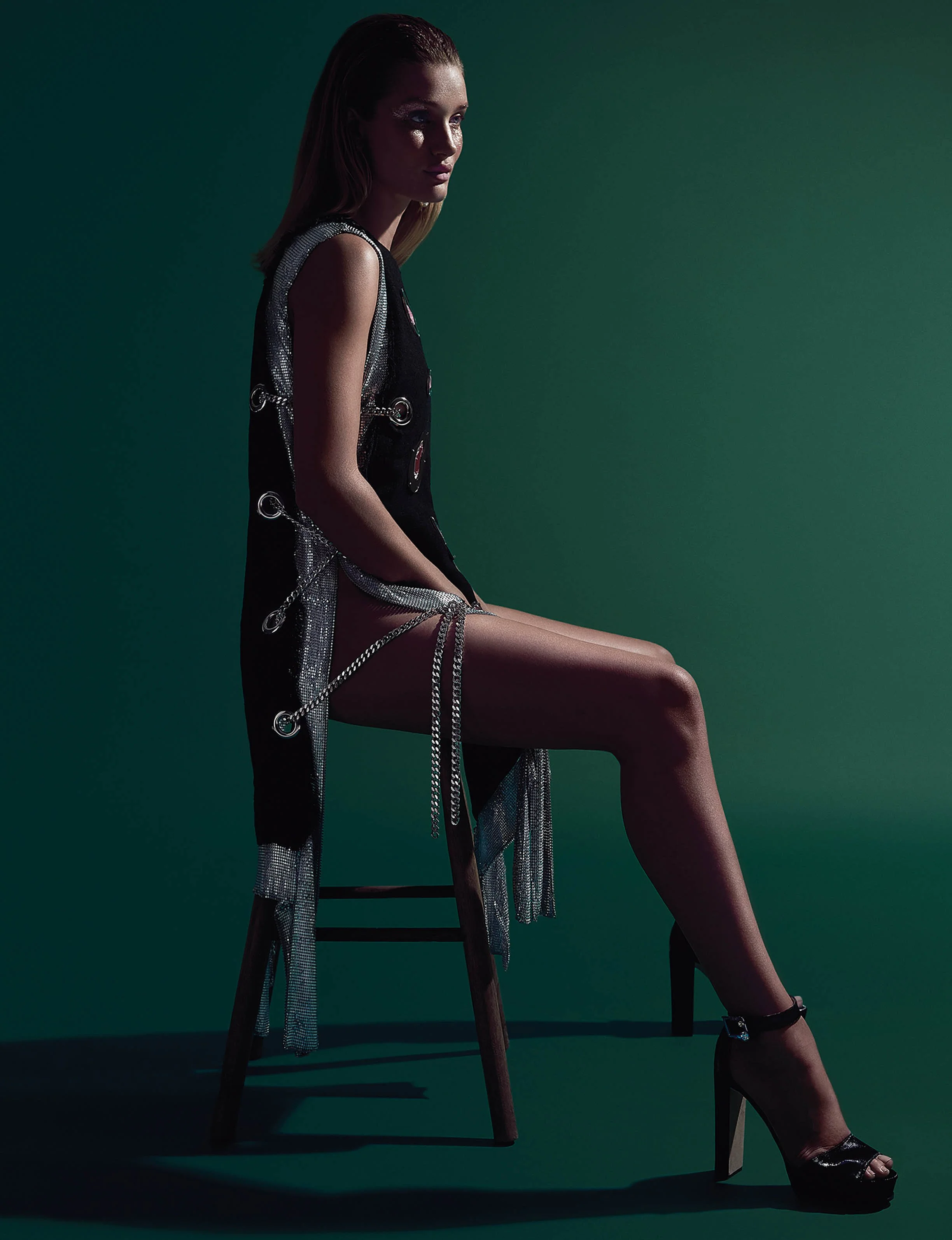 A woman sitting on a chair against a green background, wearing a black dress with silver chains and high heels.