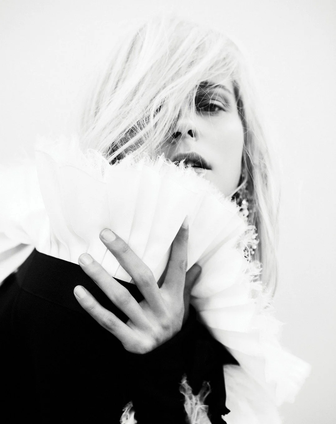 Black and white portrait of a woman with tousled hair partially covering her face, holding a white feathered fan close to her chest, with a neutral background.
