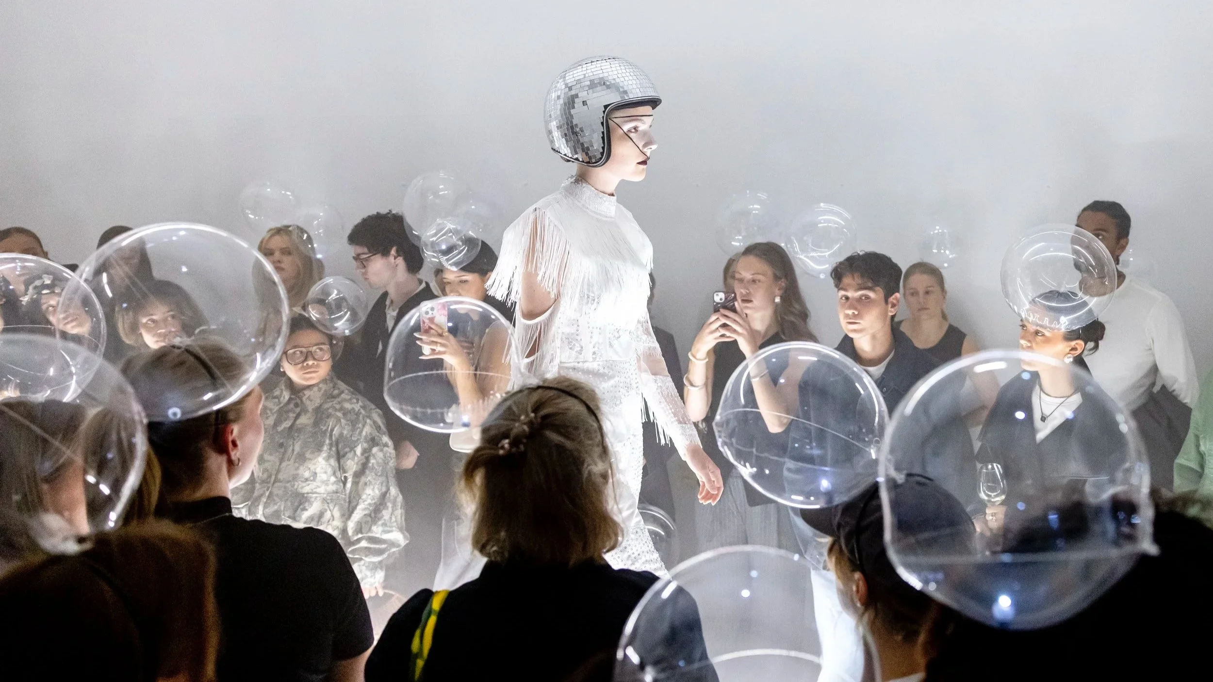 A model in a white dress and a sparkly helmet walks on a runway surrounded by audience members, some of whom are taking photos. Many audience members wear clear plastic hats, and some hold glasses of wine.