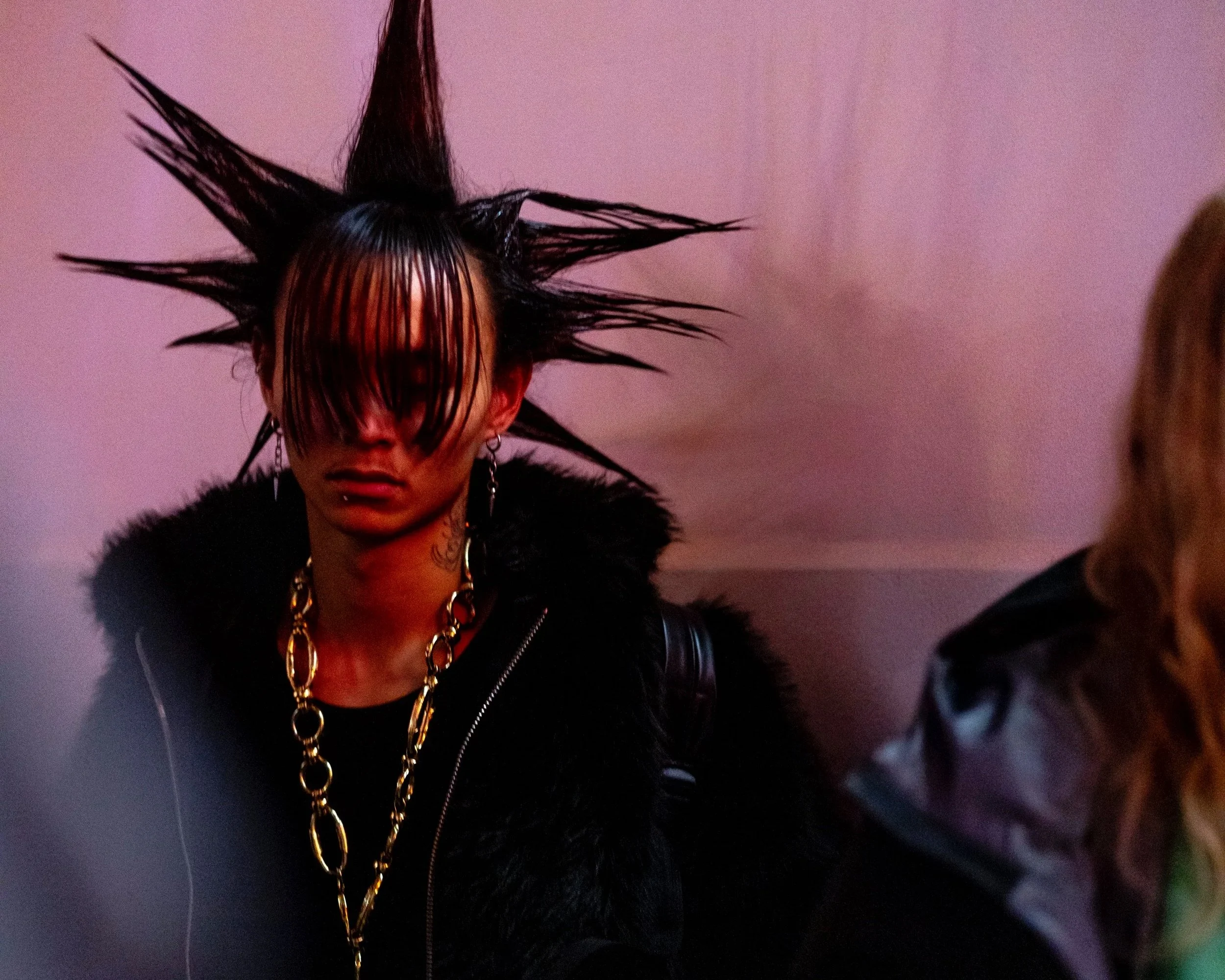 Person with spiked black hair and long bangs covering their face, wearing a black fur-lined jacket and gold chain jewelry, sitting against a pinkish wall.