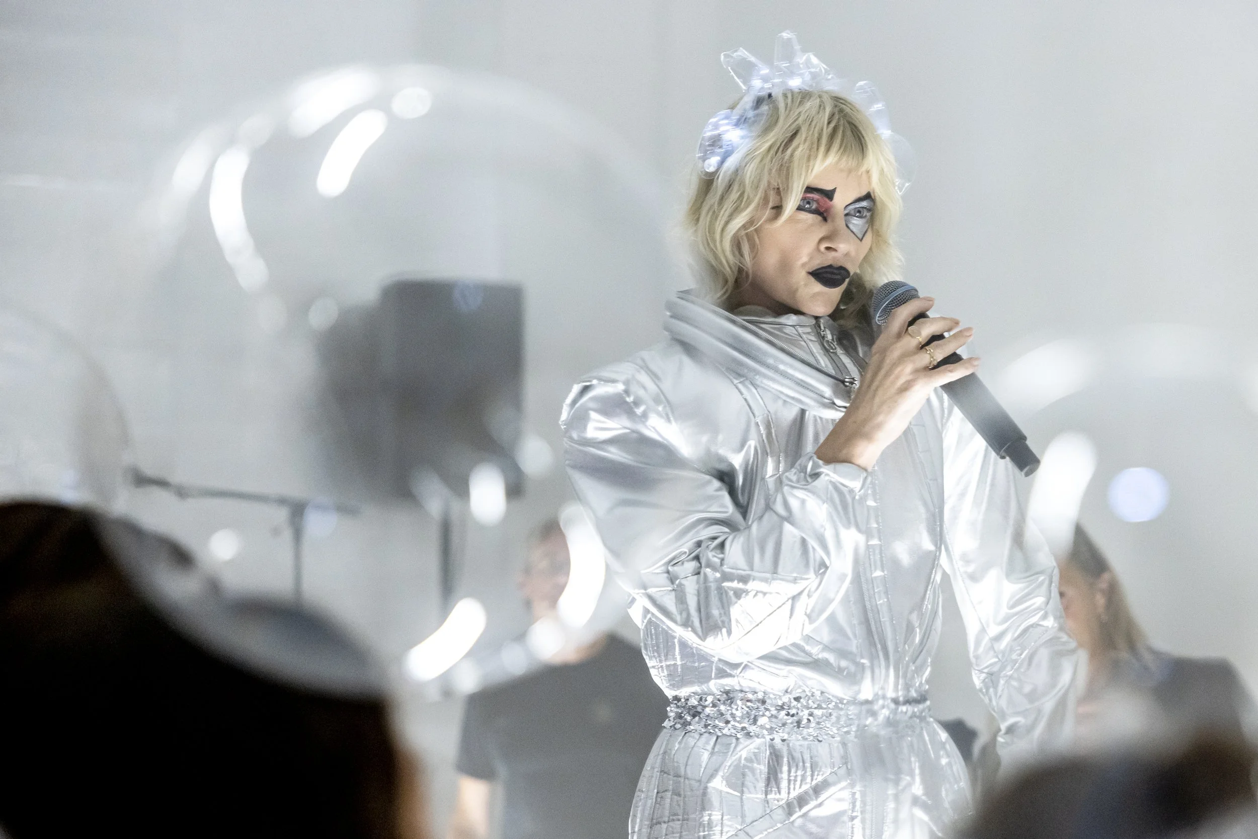 Woman dressed in a shiny silver futuristic outfit, holding a microphone, with bold makeup, blonde hair, and wearing a headpiece made of plastic, standing in front of a blurred audience.