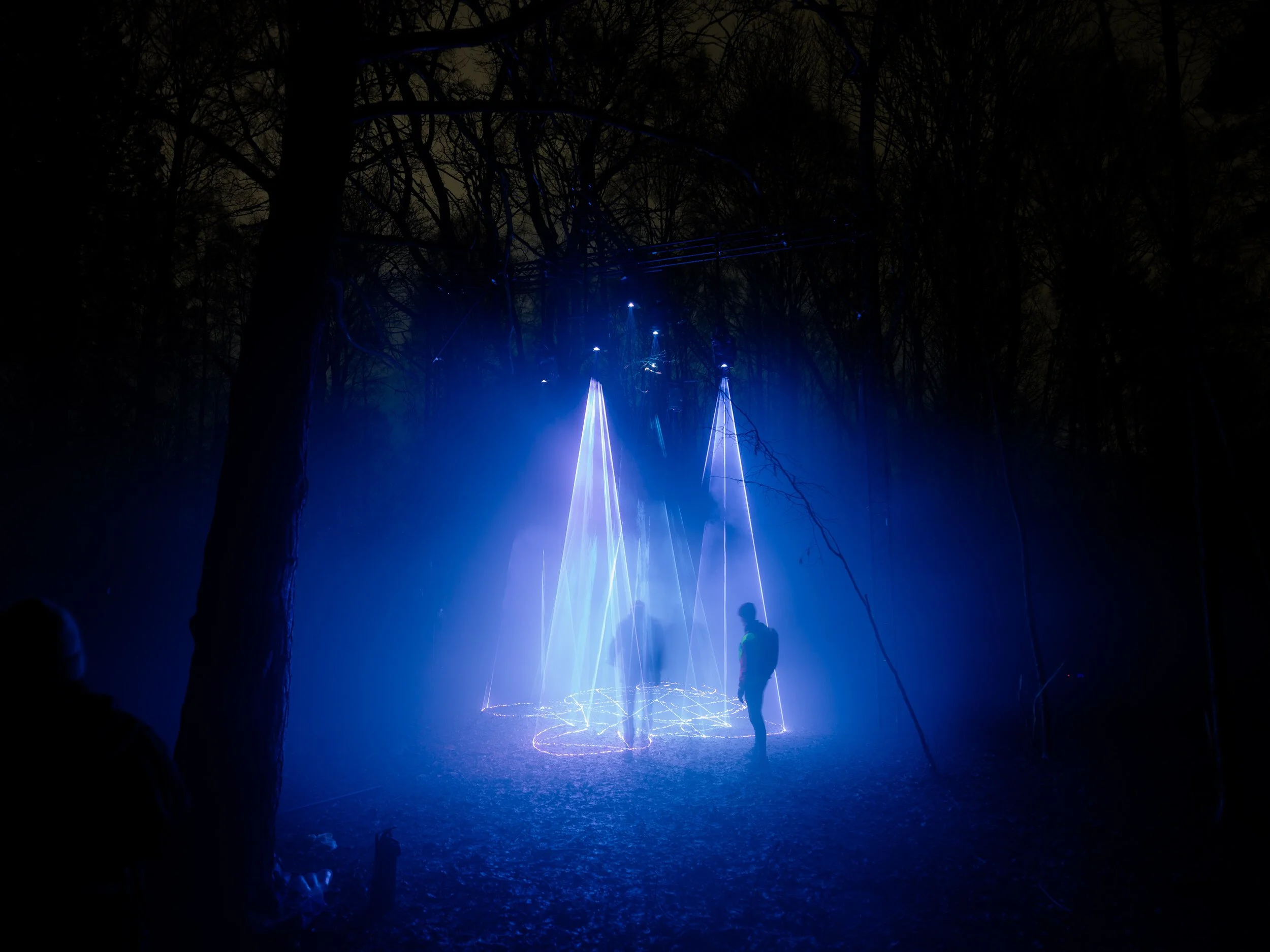 Cathedral of Ego: Light, Code, and Transcendence  MAXIME PLESCIA-BÜSCHI AND BJØRN STAAL  x COLLECTIVE OSLO Nighttime outdoor scene featuring a person interacting with blue laser light installation among trees.