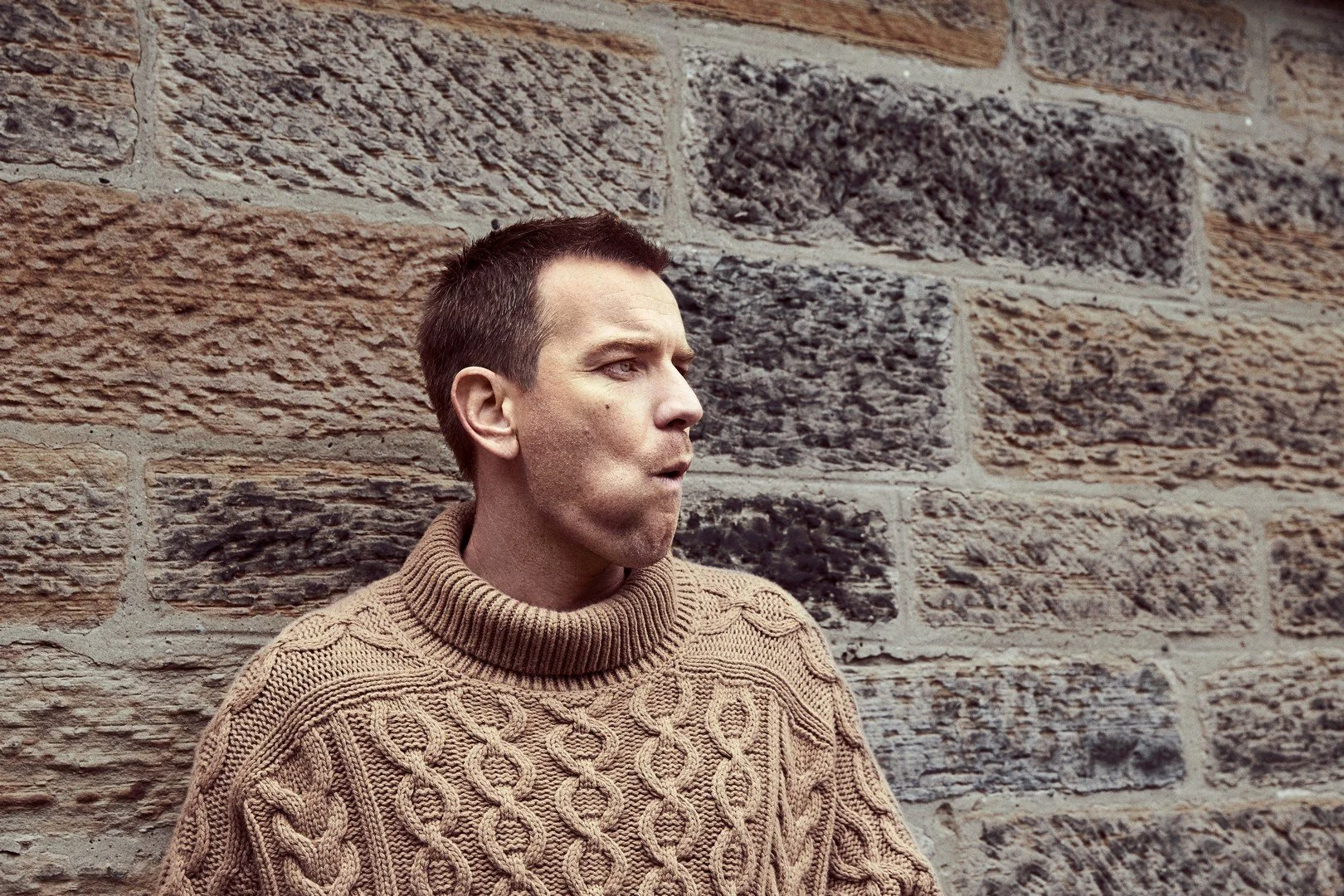 A man with short brown hair and light skin wearing a beige cable-knit sweater stands against a textured stone wall, looking to the right with a serious expression.