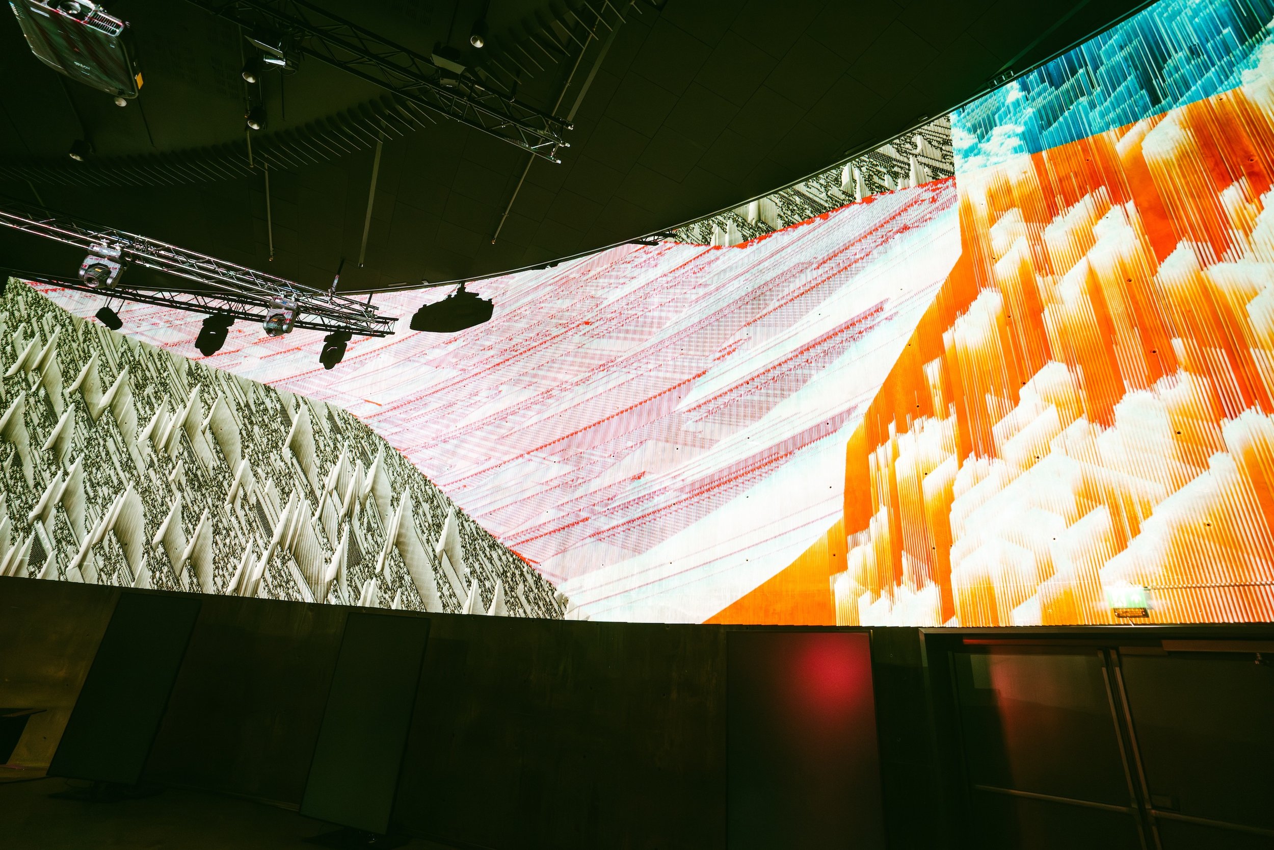 Large indoor digital screen displaying an abstract colorful image with shades of orange, white, and blue, mounted above a dark wall with stage lighting and equipment.