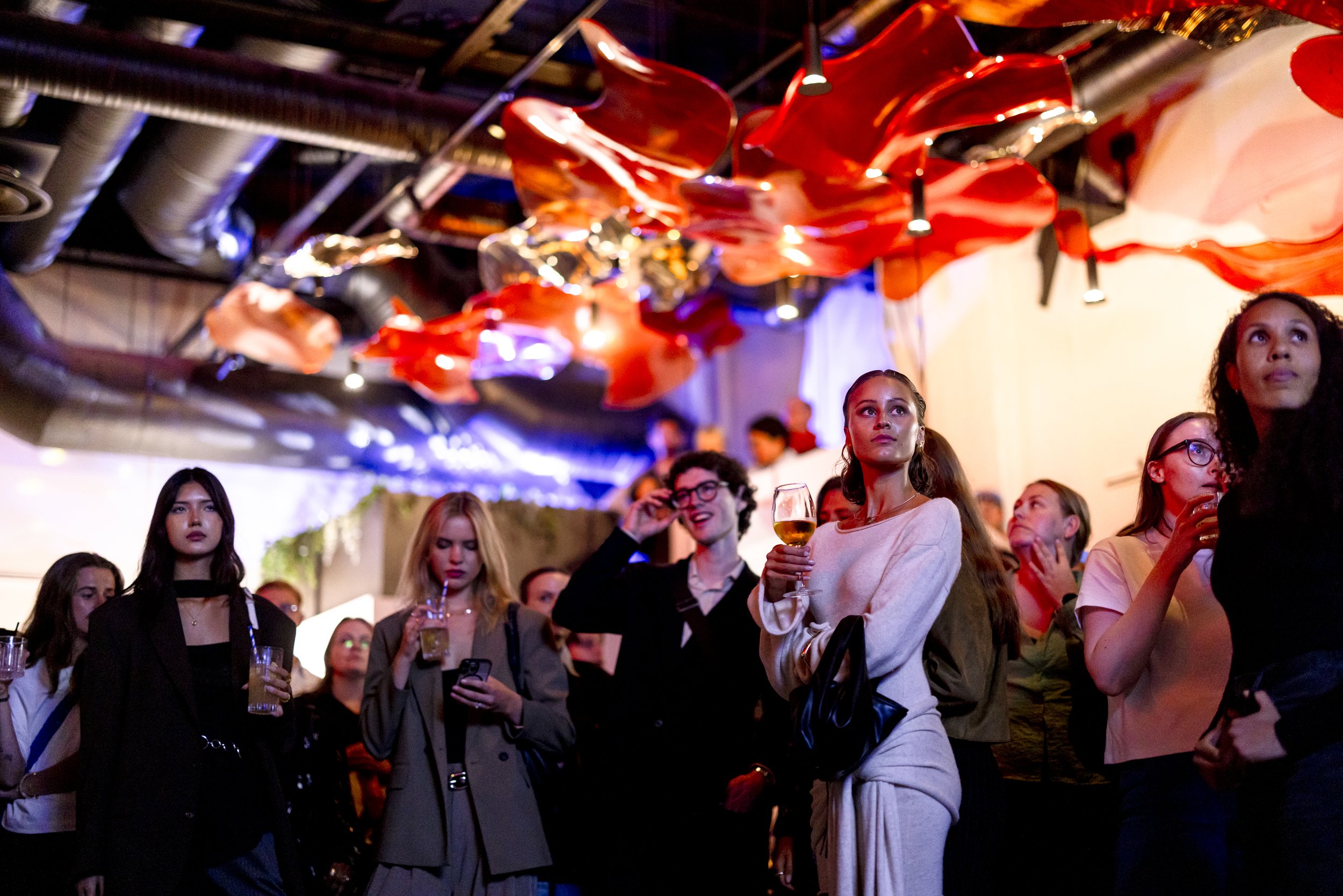 People attending a social event or party, some holding drinks, with colorful decorative ceiling elements and dim lighting.