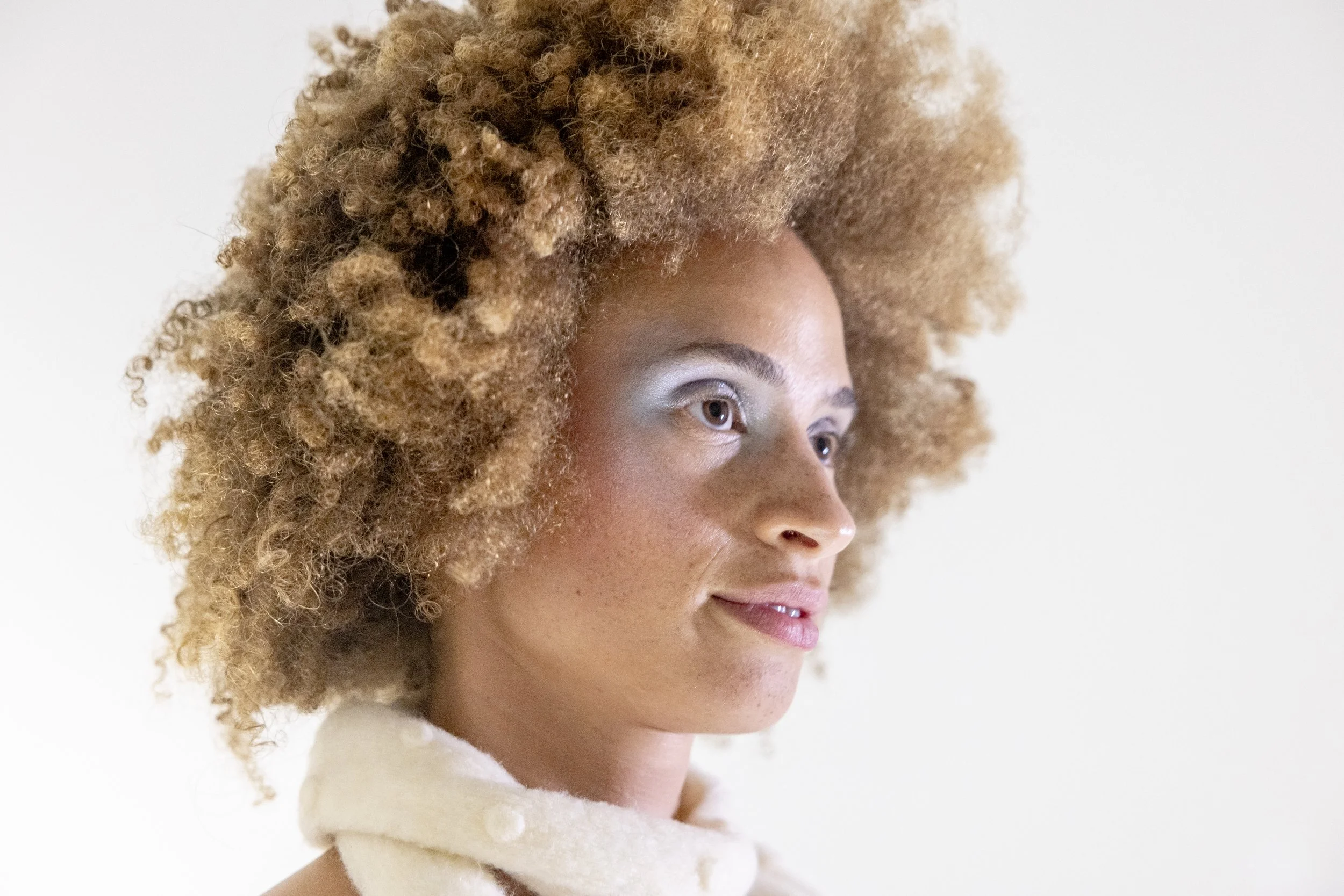 A woman with light brown skin and glowing skin, wearing a cream-colored scarf, has curly, voluminous, blonde-to-brown hair. She is looking to her left with a slight smile, against a plain white background.