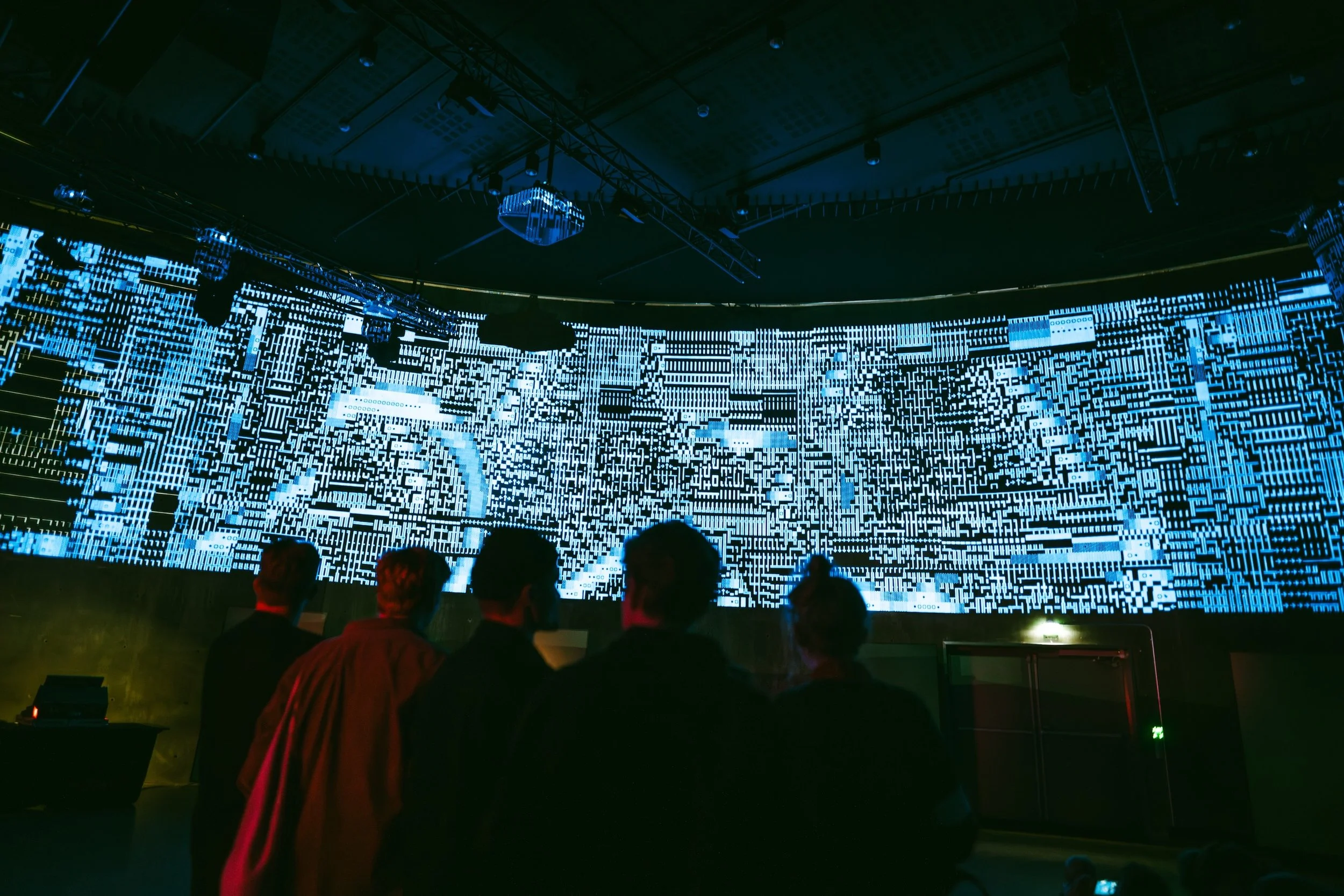 People observing a large digital display with blue and white digital patterns in a dark room