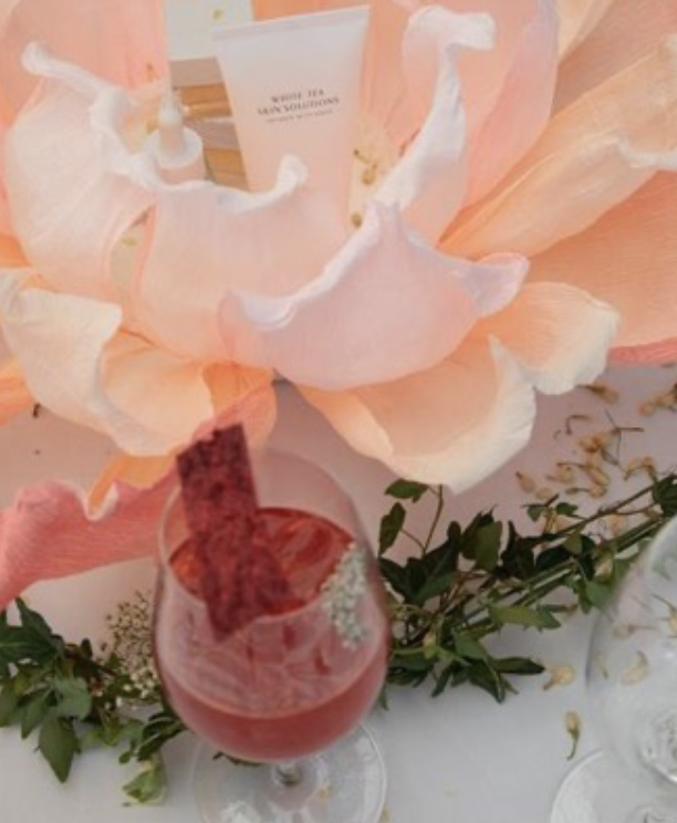 Close-up of a pink cocktail with a rectangular pink garnish on a white table, decorated with green leaves and flower petals, and a large peach-colored flower in the background.