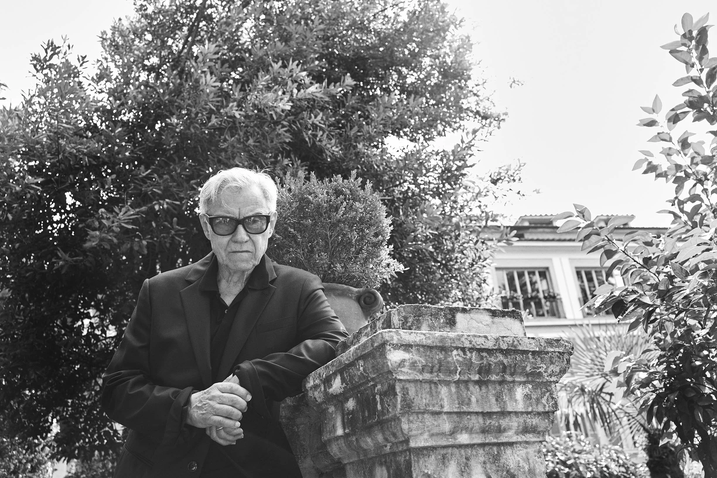 An elderly man with light-colored hair, dark glasses, and a black suit is standing outdoors next to a stone planter and a large plant. In the background are trees and a building with large windows.