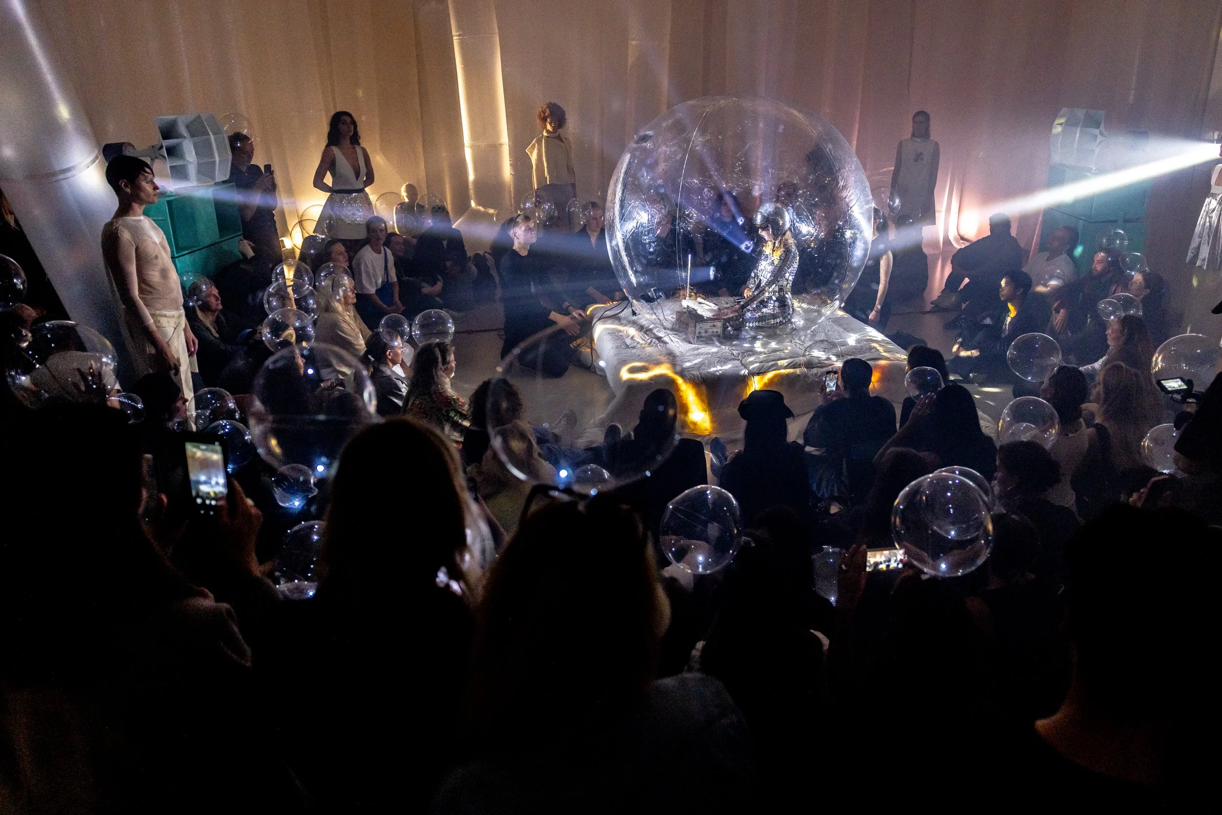 A nightclub scene with a DJ inside a large transparent bubble, surrounded by a crowd of people with many holding smartphones, and floating glass or plastic balls, creating a futuristic and immersive atmosphere.