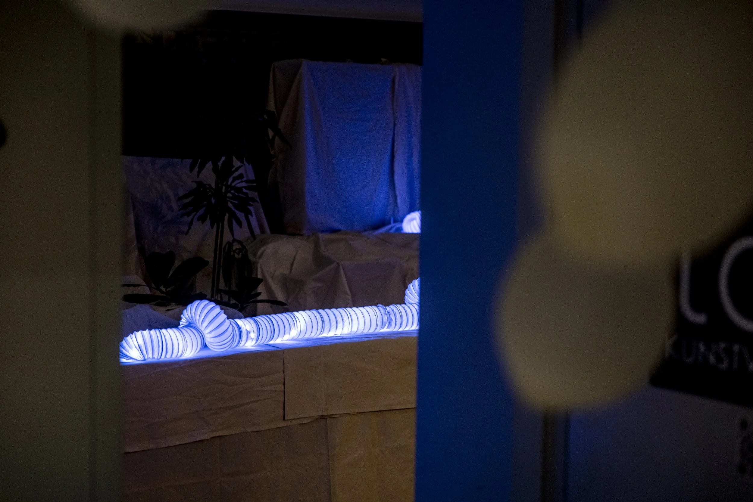 A dark room with a yellowish wall in the foreground, partially obscuring the view of a bed with a beige or light brown bedspread. On the bed, there is a glowing, flexible, ribbed light strip emitting a blue glow, arranged loosely along the edge of th