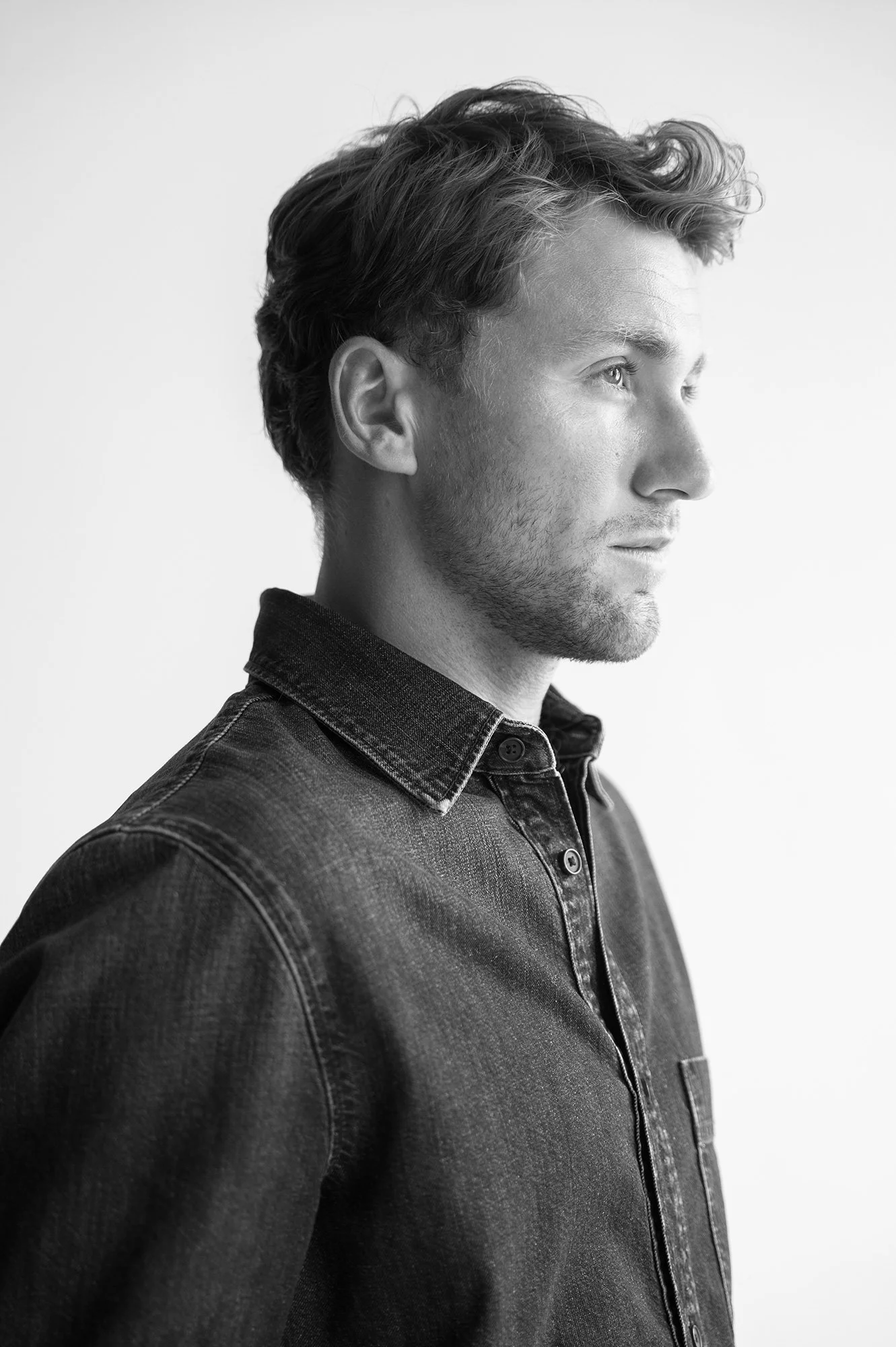 Black and white profile portrait of a young man with wavy hair wearing a denim jacket, looking to the right.
