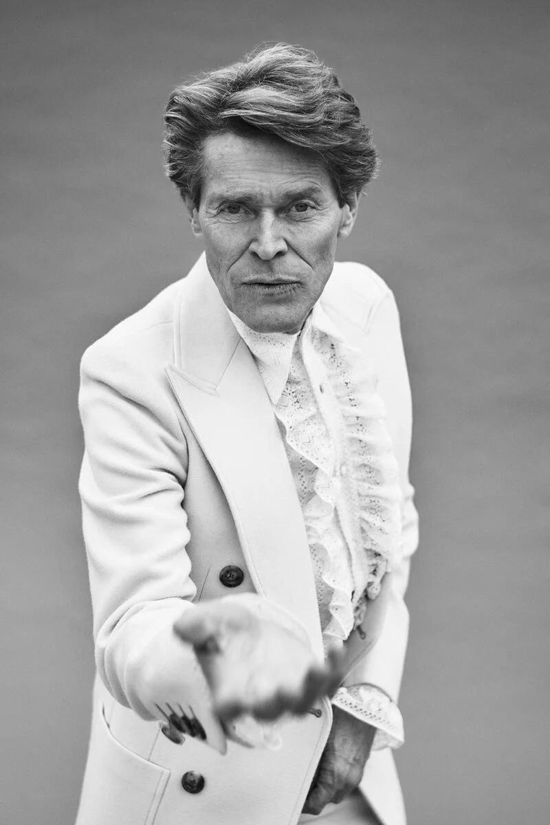 Black and white portrait of a man with styled hair, wearing a formal white suit with a ruffled shirt, pointing directly at the camera with a serious expression.