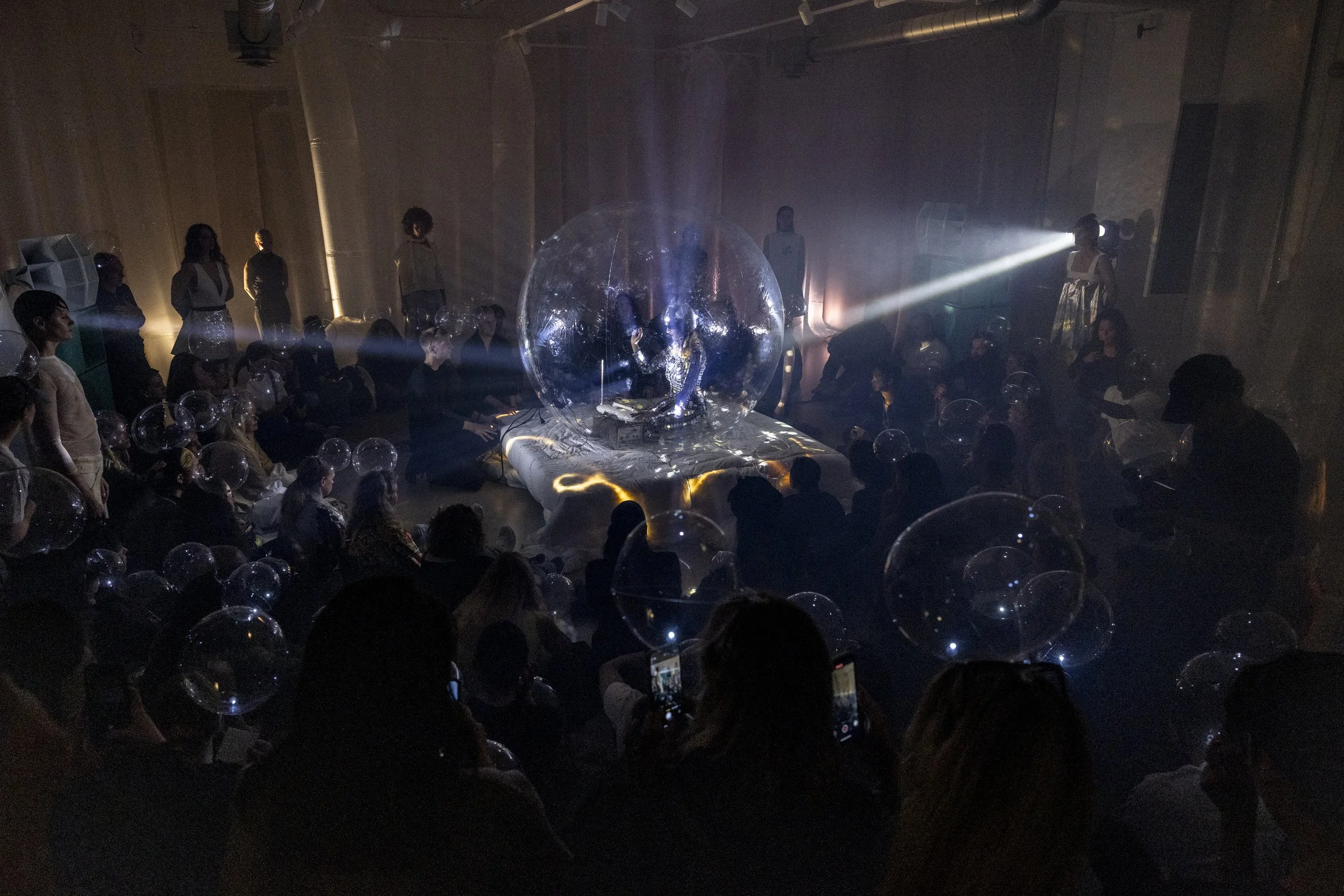 A dark room filled with people sitting and watching a performance inside a large transparent globe, surrounded by smaller transparent spheres, with dramatic lighting and spotlights highlighting the central figure.