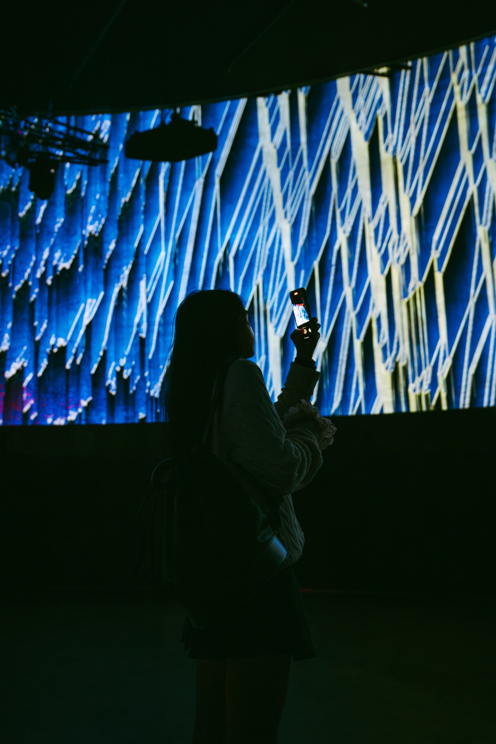 Silhouette of a woman taking a photo with her phone in front of a large digital display with vibrant blue and yellow abstract patterns.