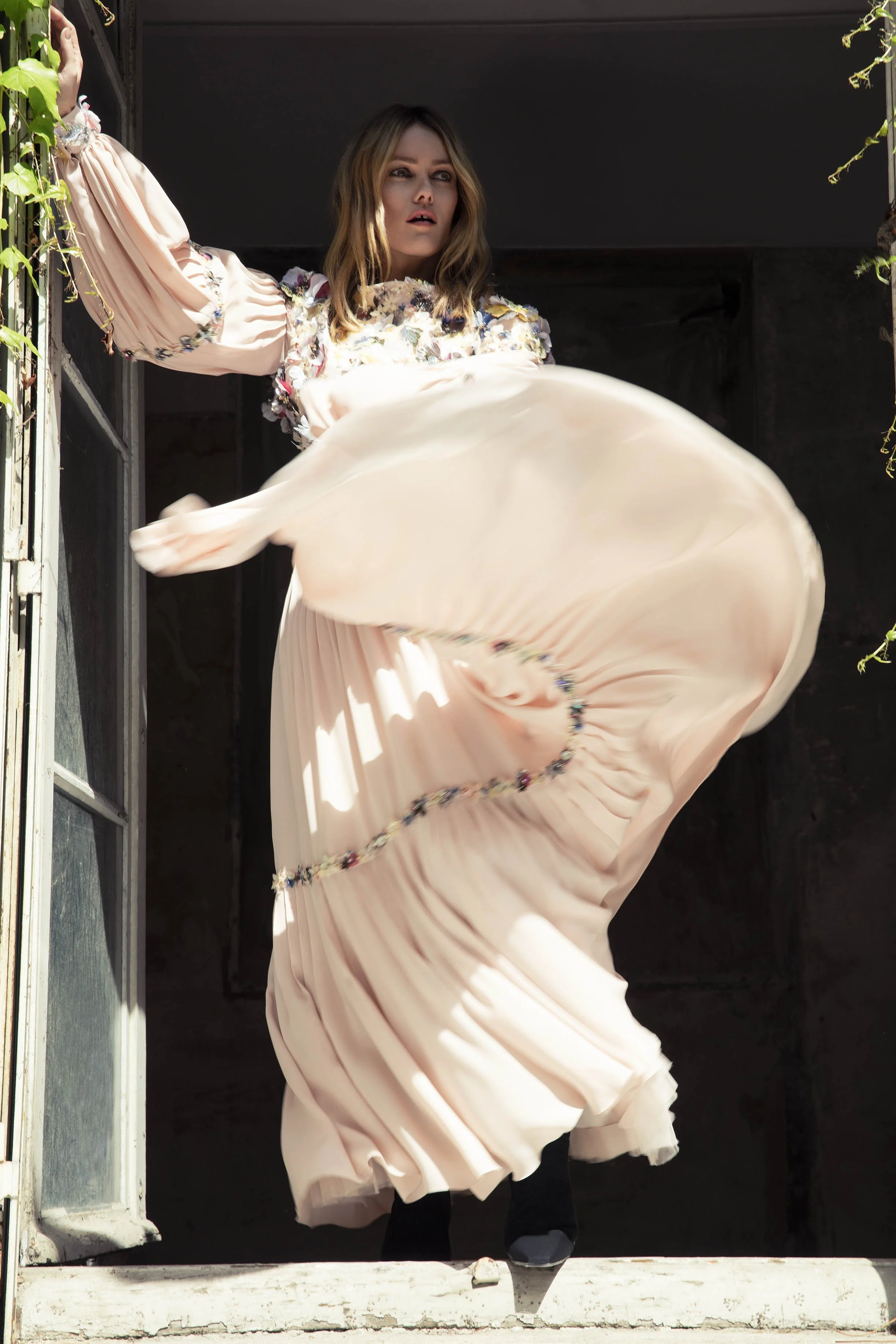 A woman in a flowing pink dress with floral embroidery, standing at a doorway with one arm raised and holding onto the doorframe, as her skirt billows outward.