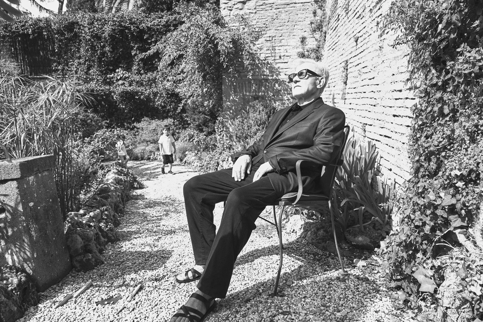A man wearing sunglasses, a black blazer, and black pants sitting on a metal chair in a garden with two children in the background.