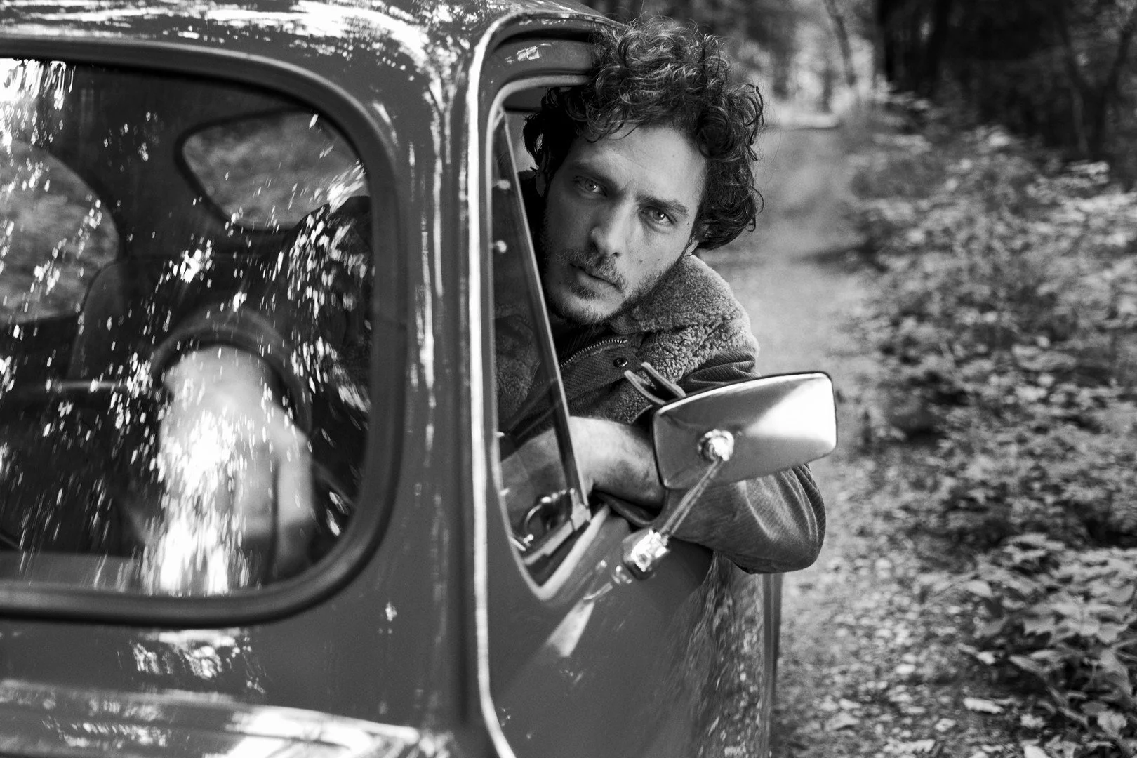 Black and white photo of a man with curly hair leaning out of a vintage car window, looking directly at the camera, on a wooded road.