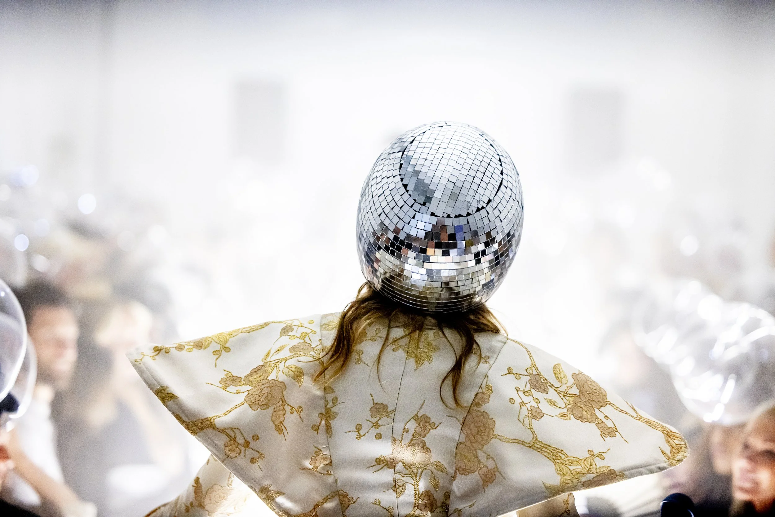 Person at a party wearing a disco ball helmet and a white floral kimono-style garment, with blurred other partygoers in the background.