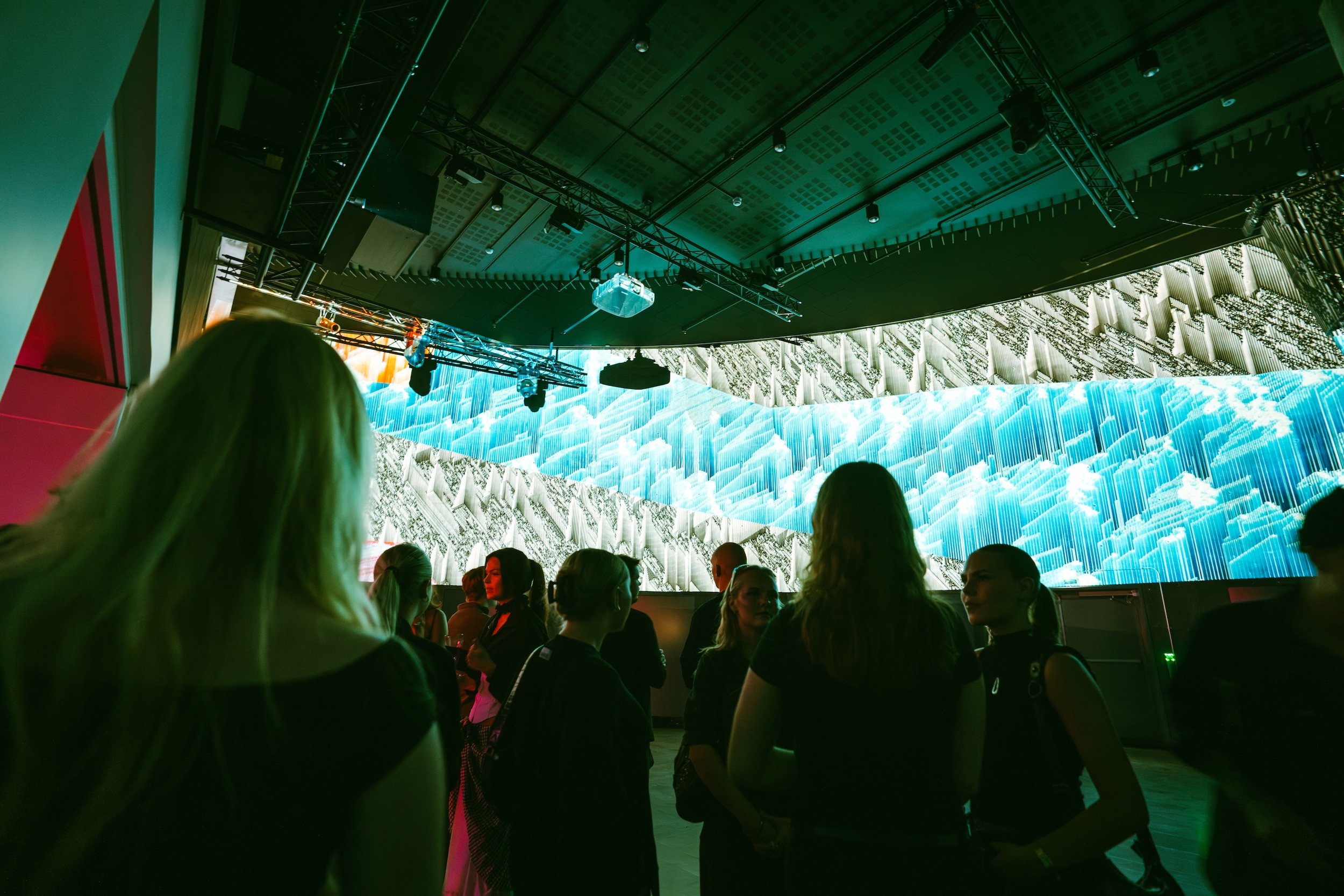 People gathered in a dark room with an illuminated large digital screen displaying a digital landscape of clouds and mountains.