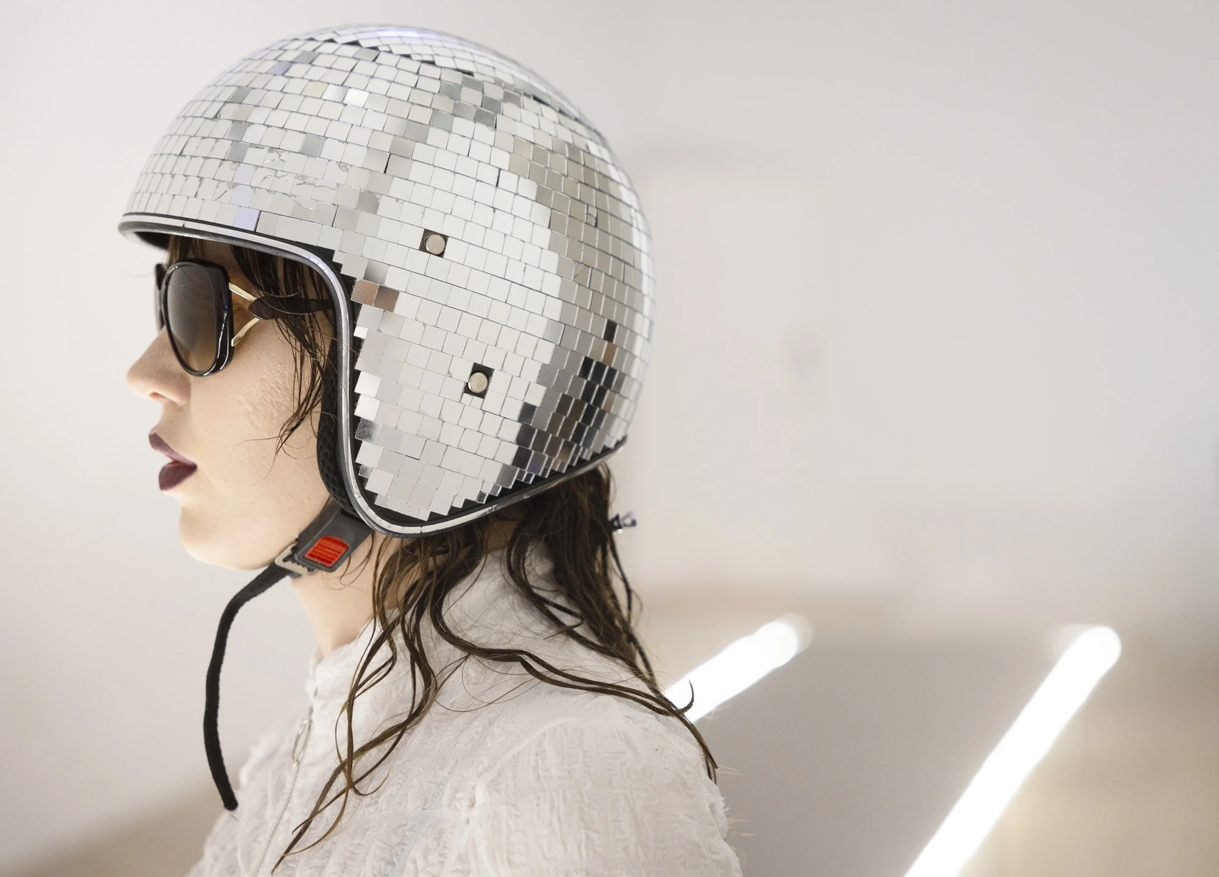 A woman wearing a mirror mosaic helmet and sunglasses, facing left, with a white textured shirt and wavy hair.
