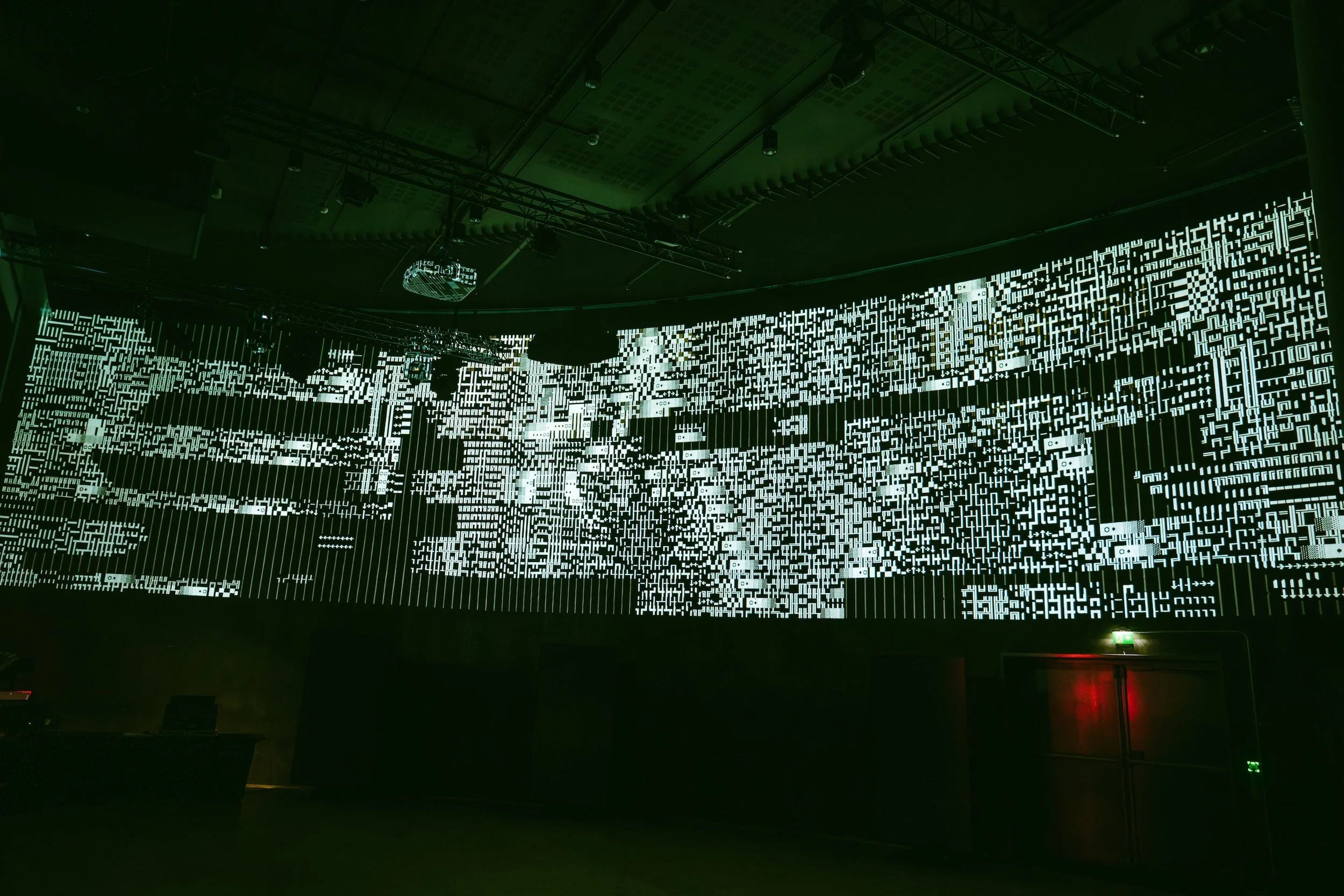 Large digital display with white abstract patterns and symbols on a dark background, in a dimly lit room or auditorium.