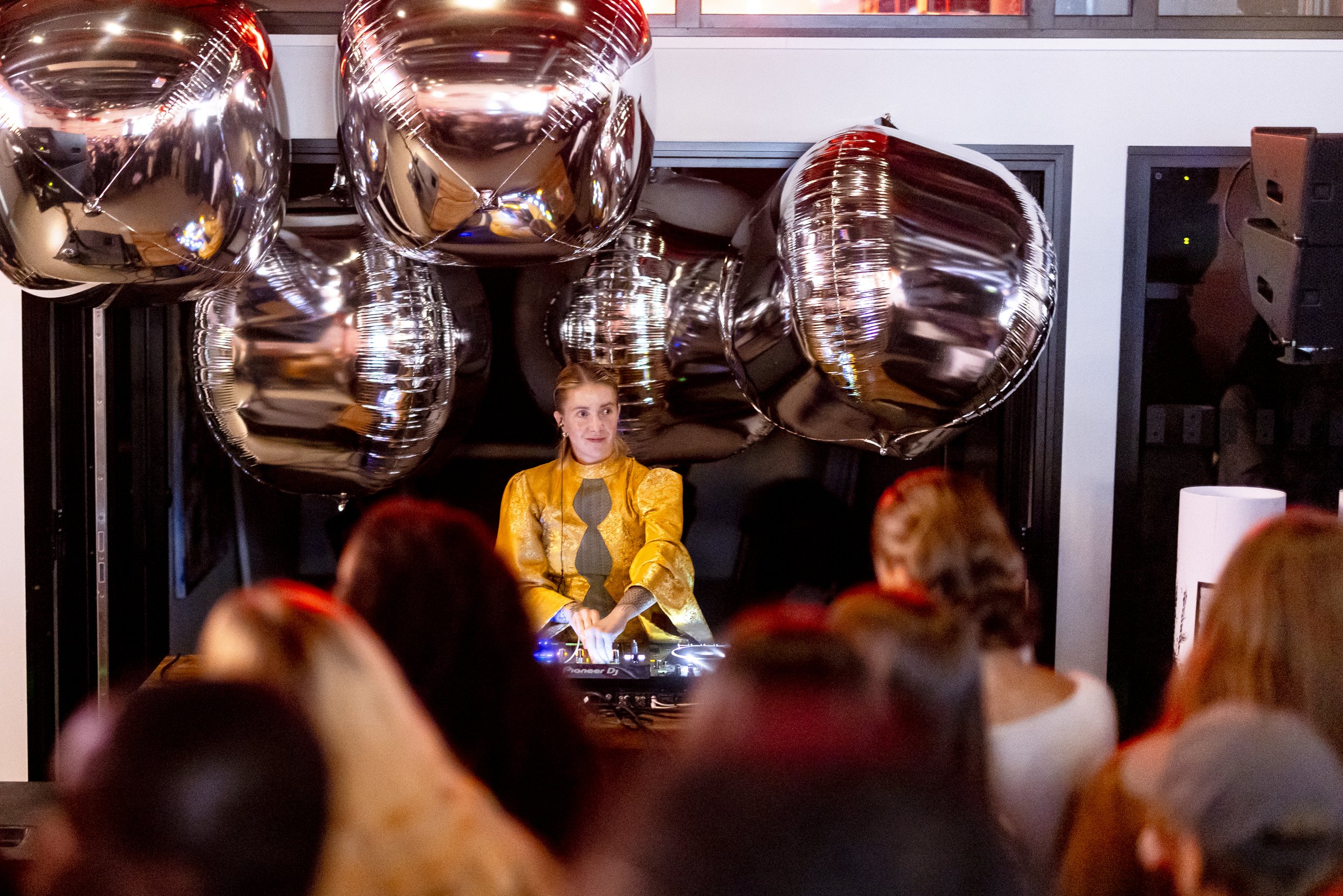 A DJ wearing a yellow and gold patterned jacket performing at a party or event with balloons in the background and an audience watching.