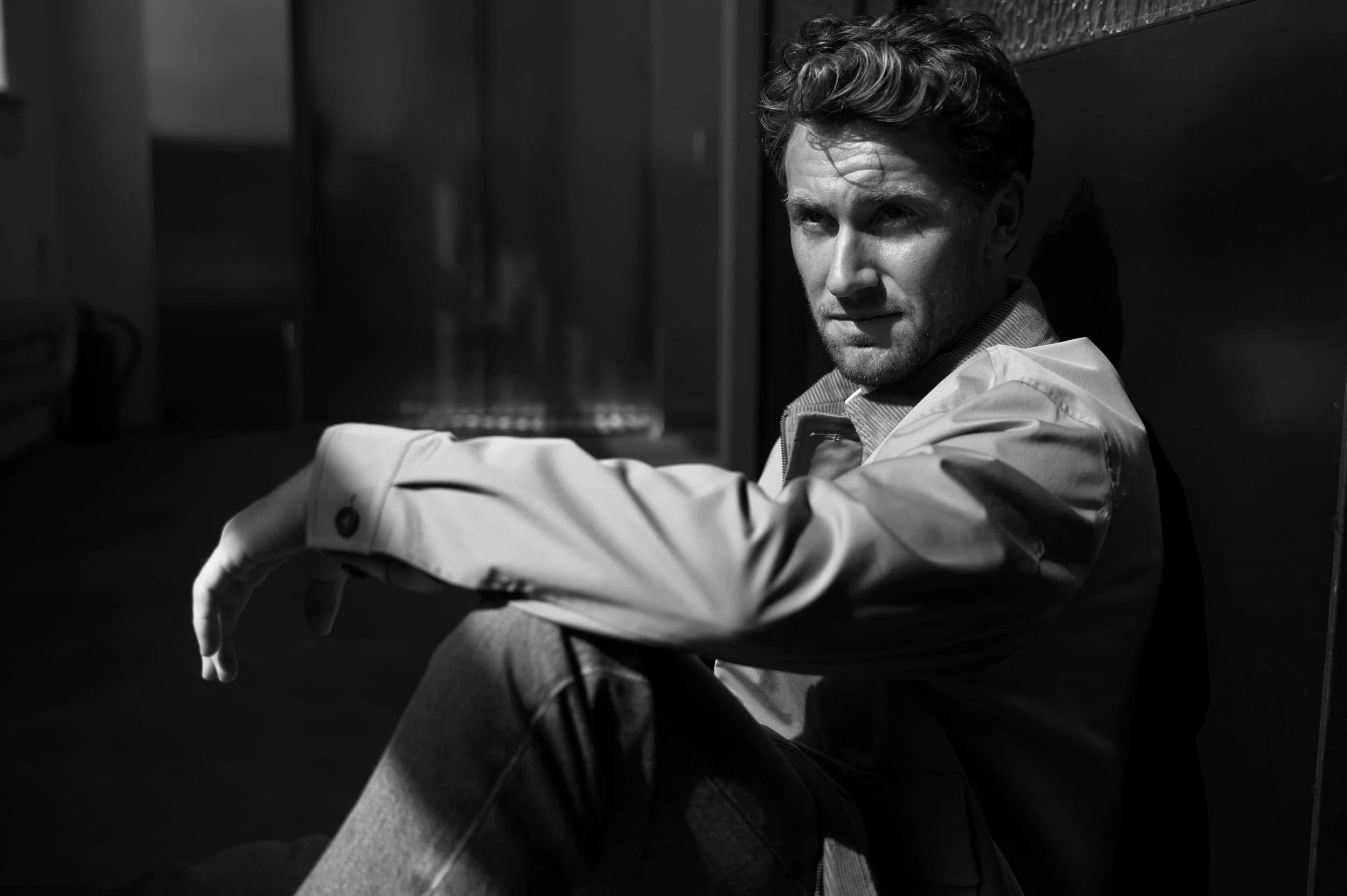 Black and white photo of a young man sitting on the floor against a wall, looking intently at the camera with a serious expression, wearing a light-colored jacket and dark pants.