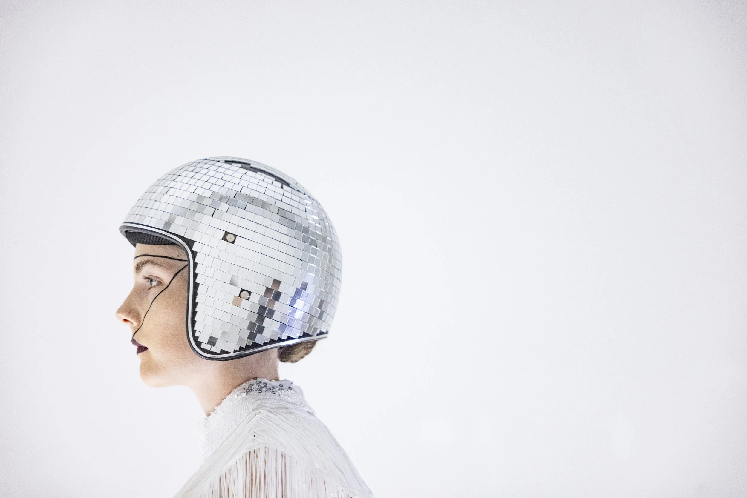 Woman wearing a shiny, mirrored disco ball helmet, standing in profile against a plain white background.