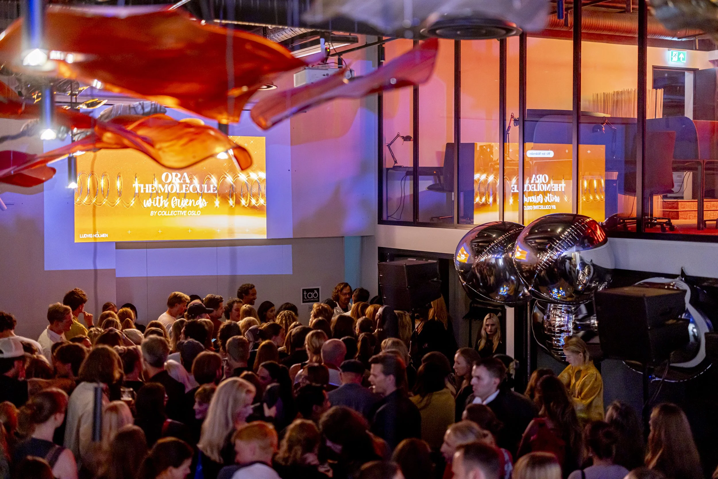 A large crowd of people gathered in a modern indoor venue for a presentation or event. There is a brightly lit screen displaying the title 'ORA THE MOLECULE with friends by COLLECTIVE OSLO' and some decorative large metallic balloons are visible. The