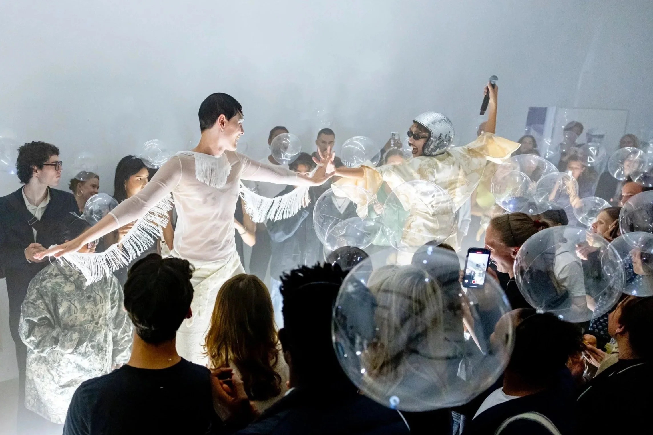 People dancing at a celebration surrounded by transparent balloons, with a woman taking photos and a man wearing a helmet and microphone.