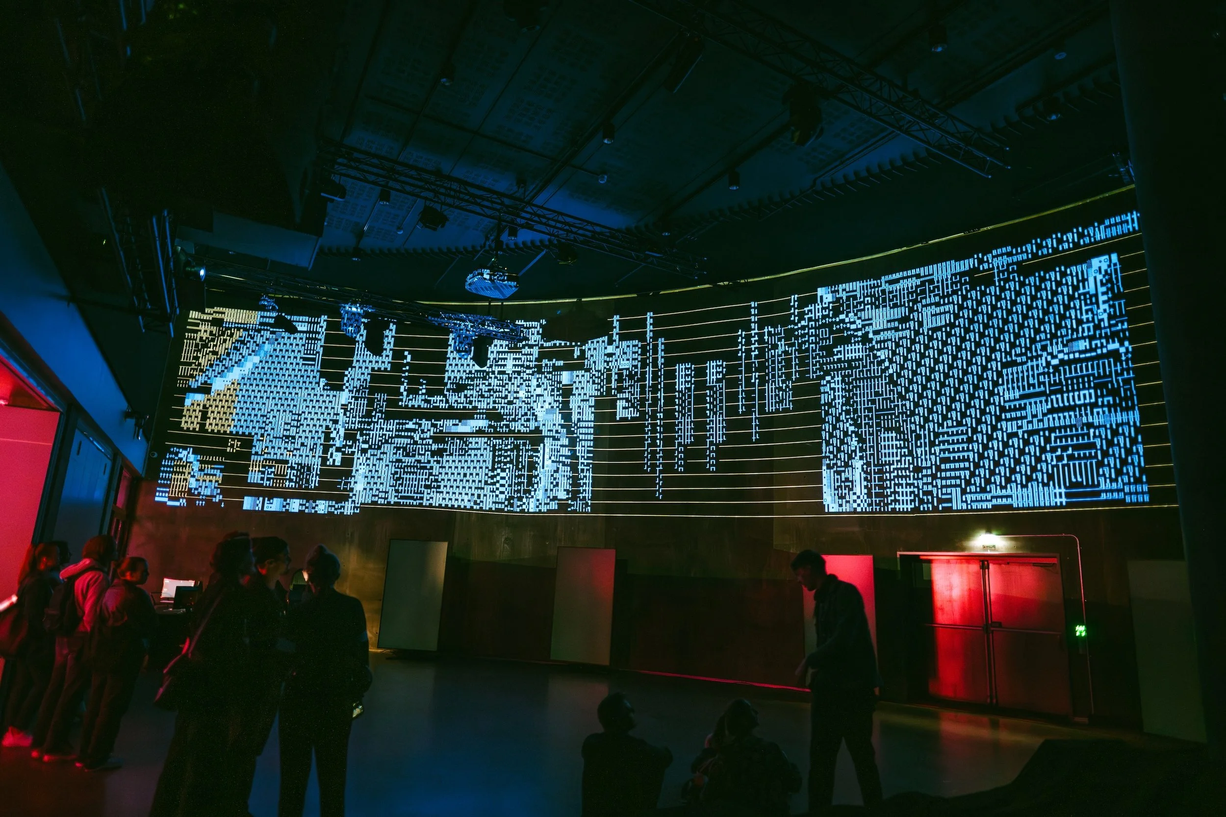 Group of people standing and sitting in front of a digital art installation featuring a glowing cityscape with intricate blue and yellow pixelated patterns on a large curved screen in a dark room.