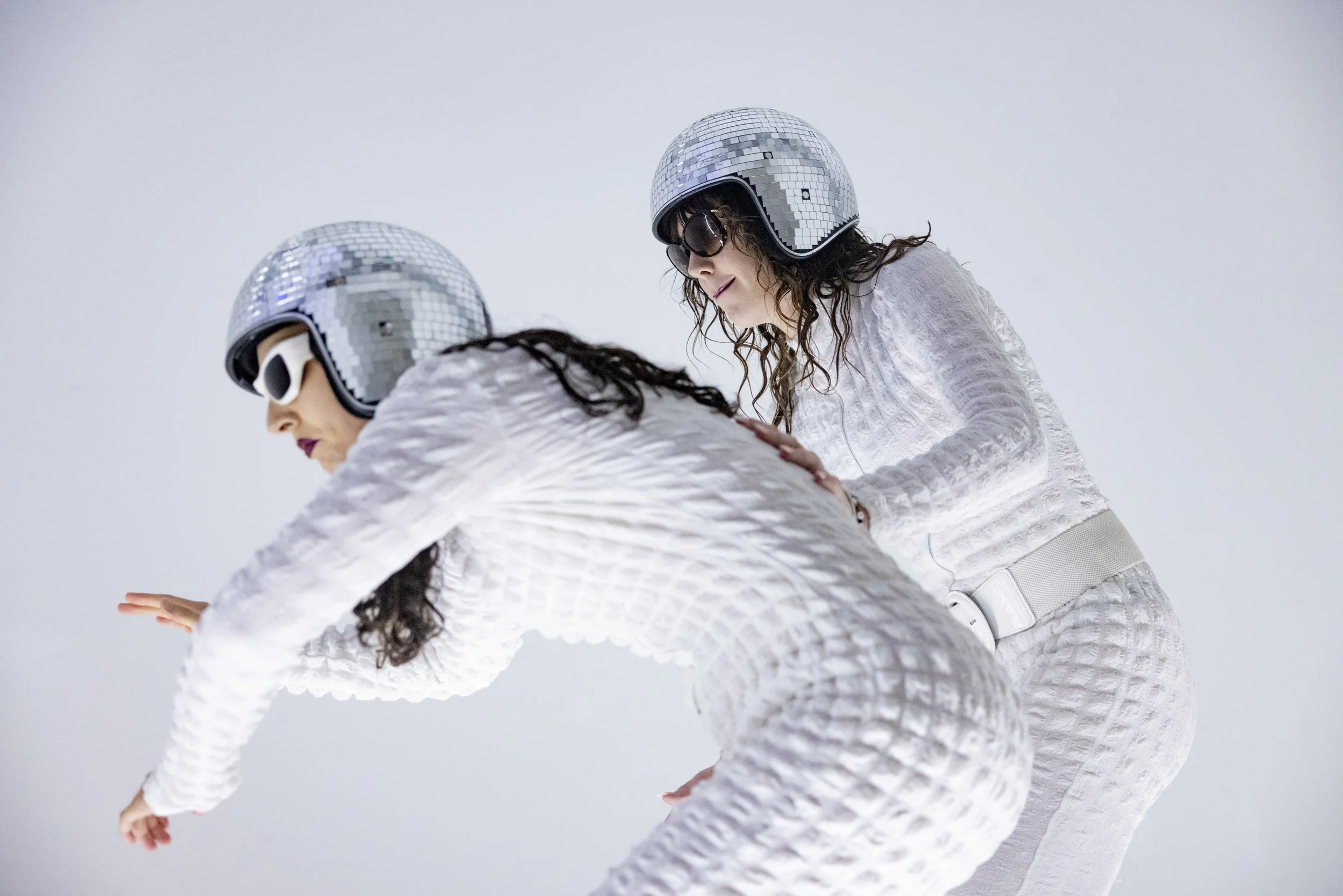 Two women wearing matching white textured outfits, mirrored disco helmets, and sunglasses, with one leaning forward as the other helps them maintain balance.