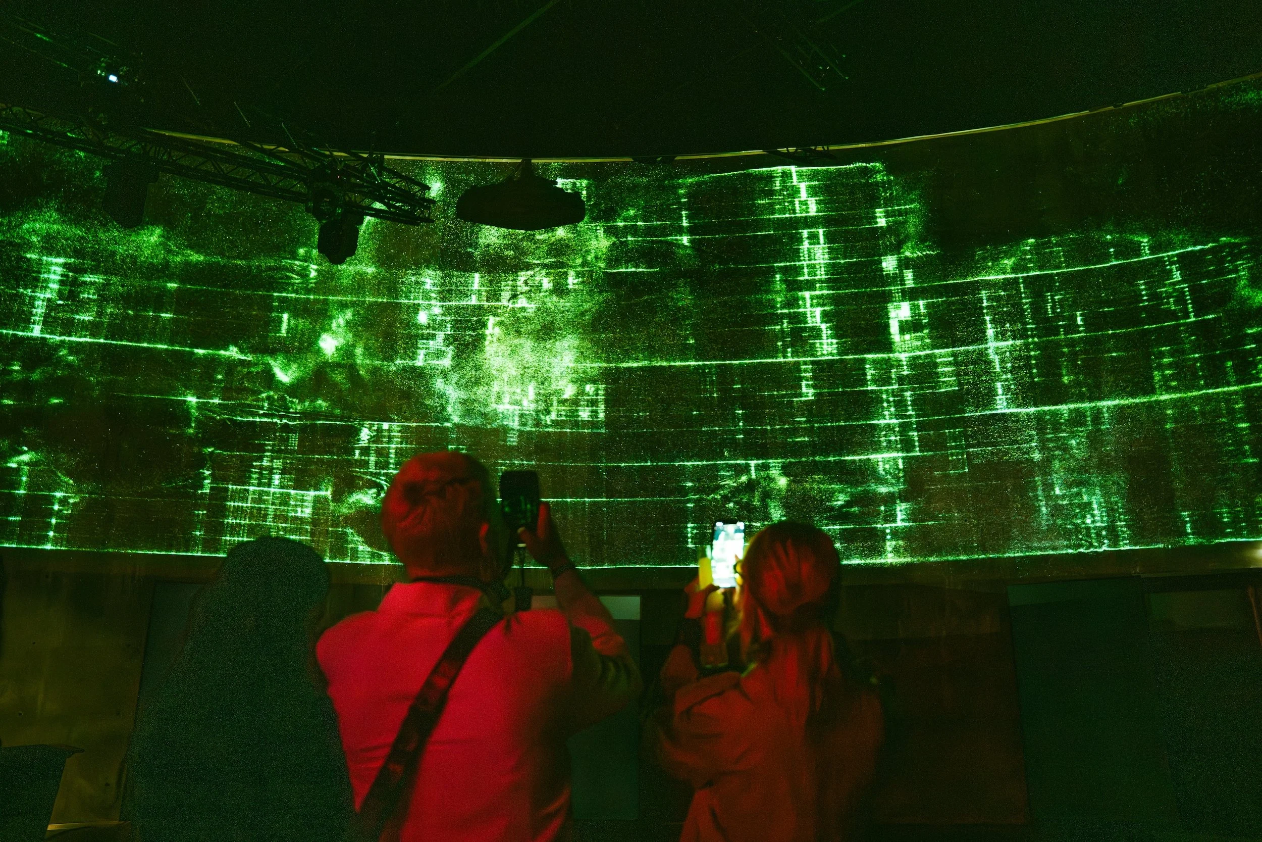 Three people are watching a digital green light display on a large curved screen in a dark environment, possibly a concert or event.
