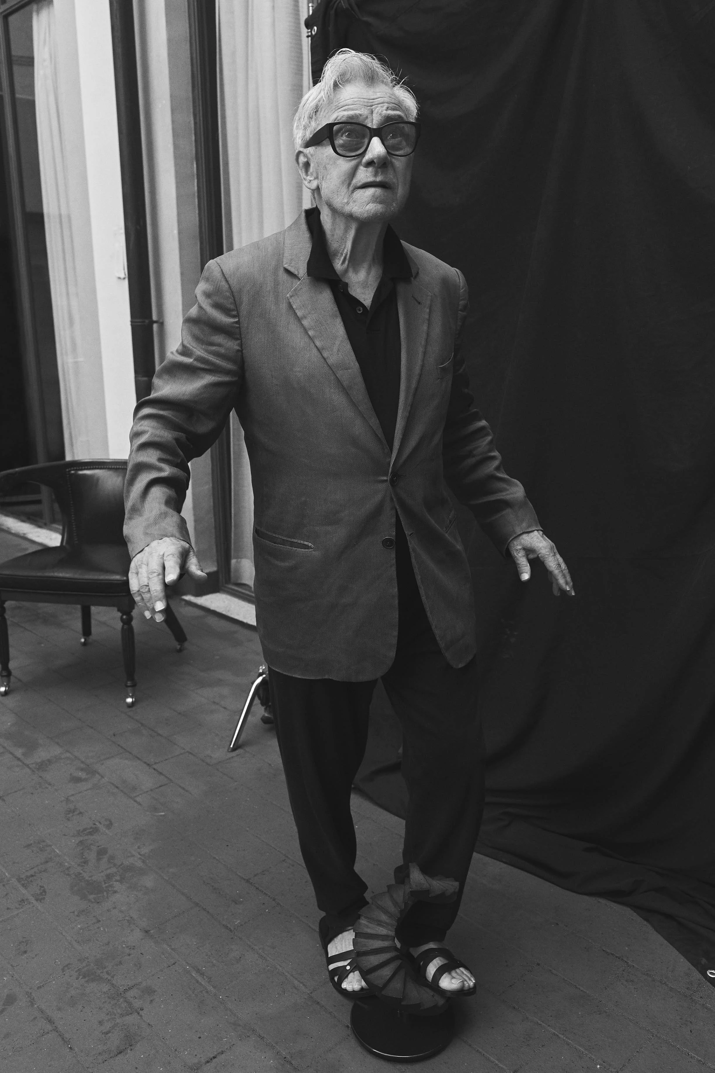 An elderly man wearing a blazer, black shirt, glasses, and sandals balances on a small skateboard.