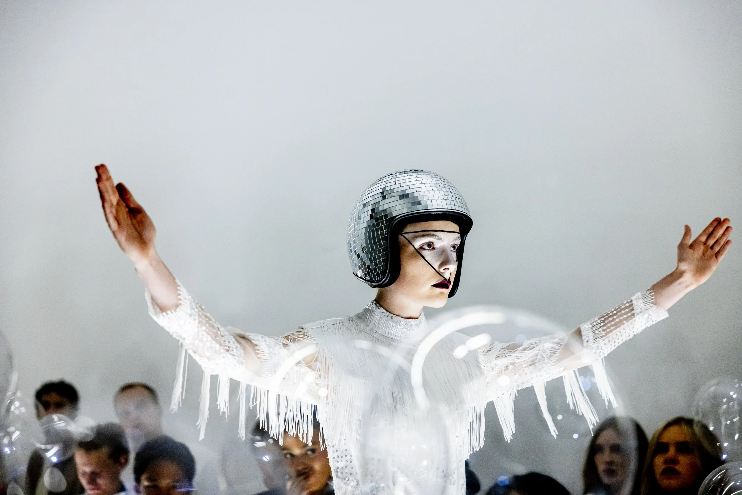 A woman wearing a metallic disco ball helmet and a white fringed outfit, with arms outstretched, surrounded by balloons, in a modern indoor setting.