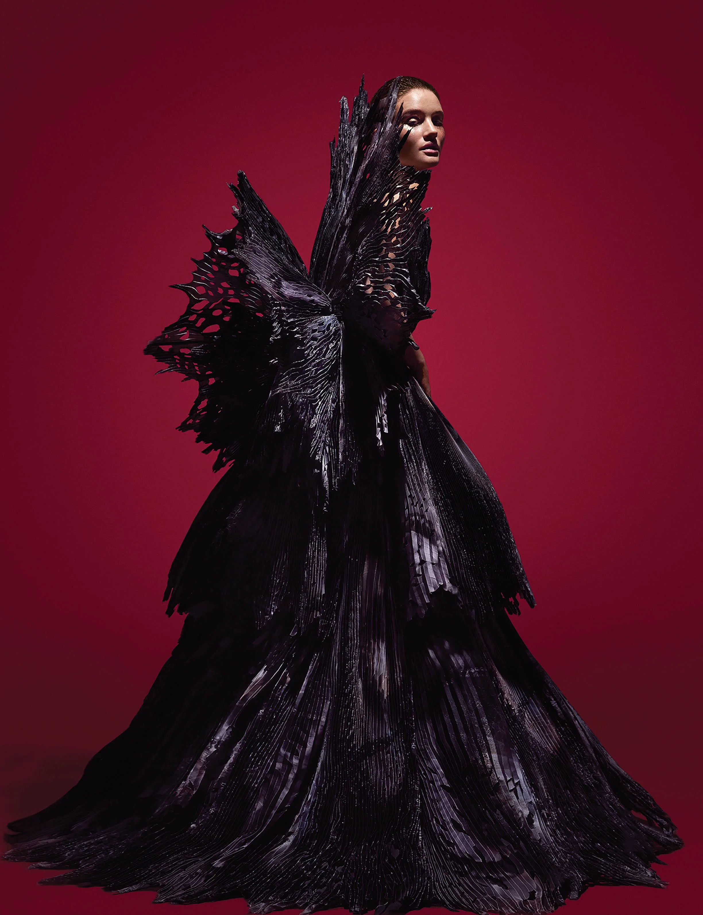 A woman in a dramatic black gown with large feathered and textured details, standing against a red background.