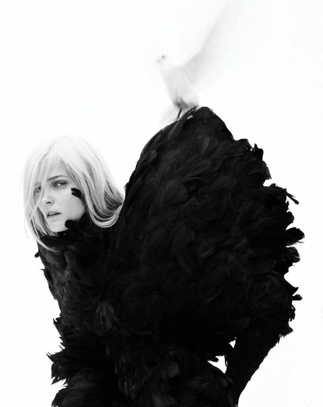 Black and white photo of a woman with blonde hair, wearing a large black feathered garment, looking over her shoulder.