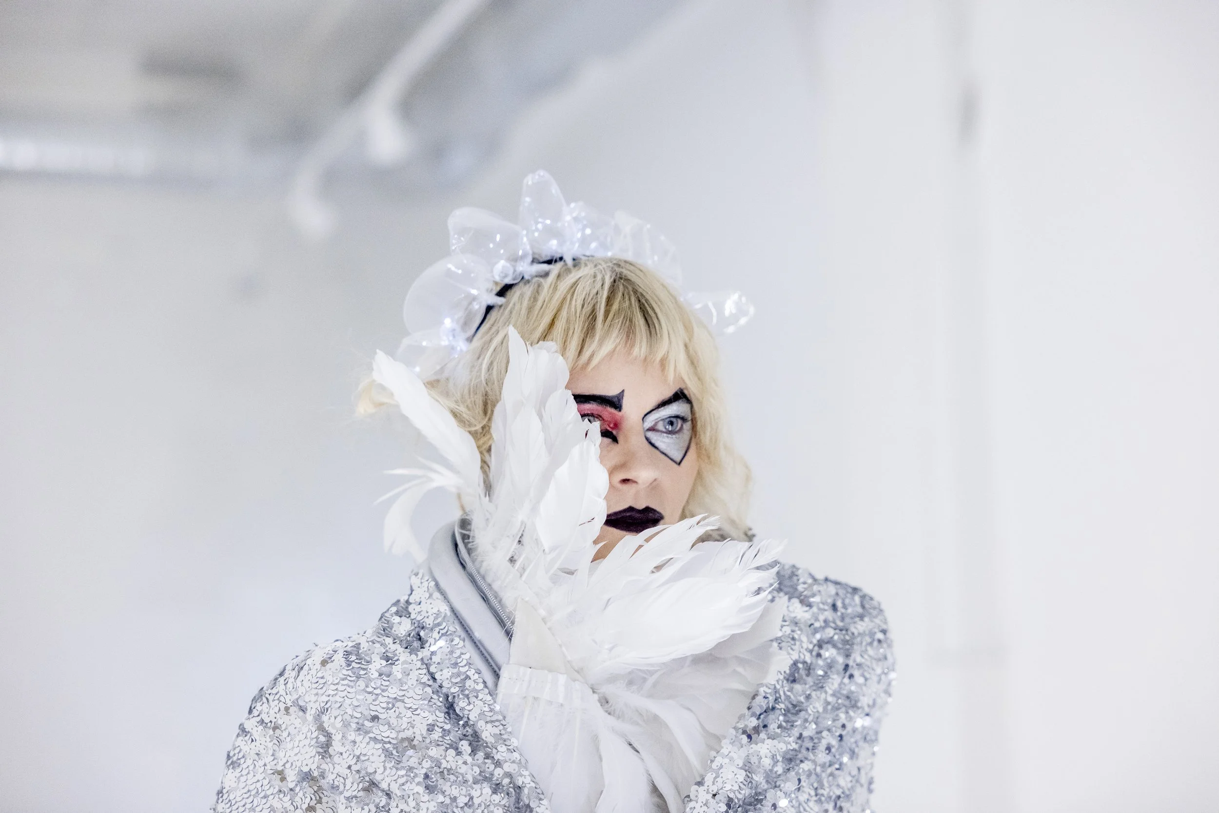 Woman with blonde hair in artistic makeup, wearing a silver sequin outfit and white feather gloves, holding a feather to her face, and wearing a transparent headpiece with LED lights.