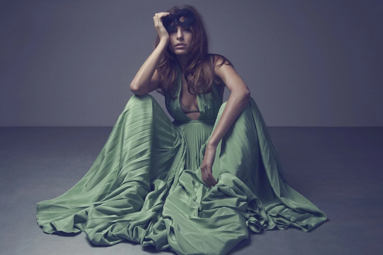 Woman sitting on the floor wearing a flowing green dress, holding a black mask to her face, with a neutral gray background.