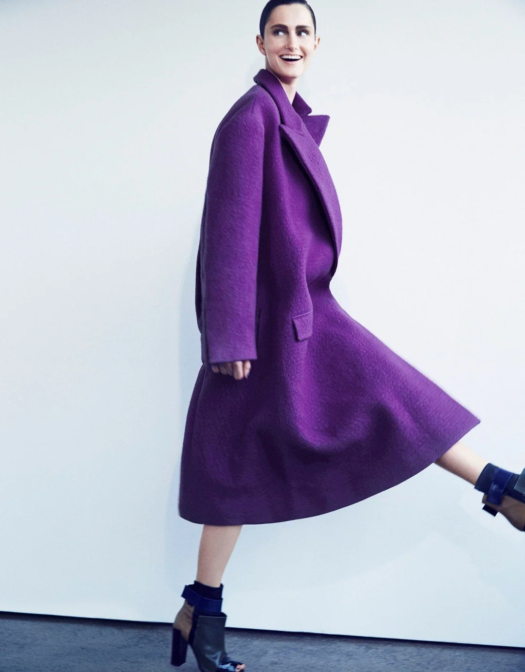 Woman smiling, wearing a purple coat and matching skirt, black heeled sandals, standing against a plain white background.